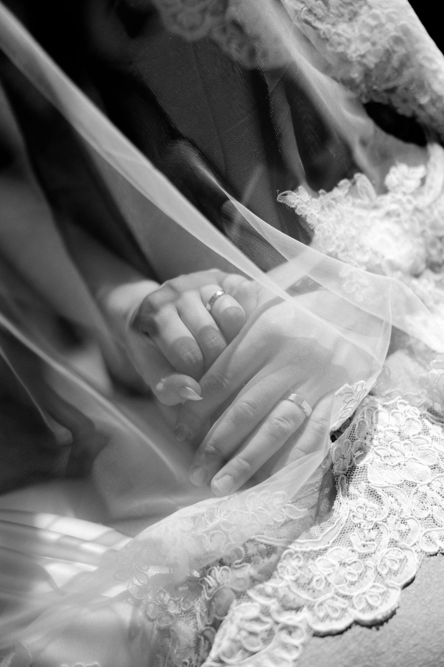 Black and white close-up of newlyweds holding hands with wedding rings visible beneath bridal veil.