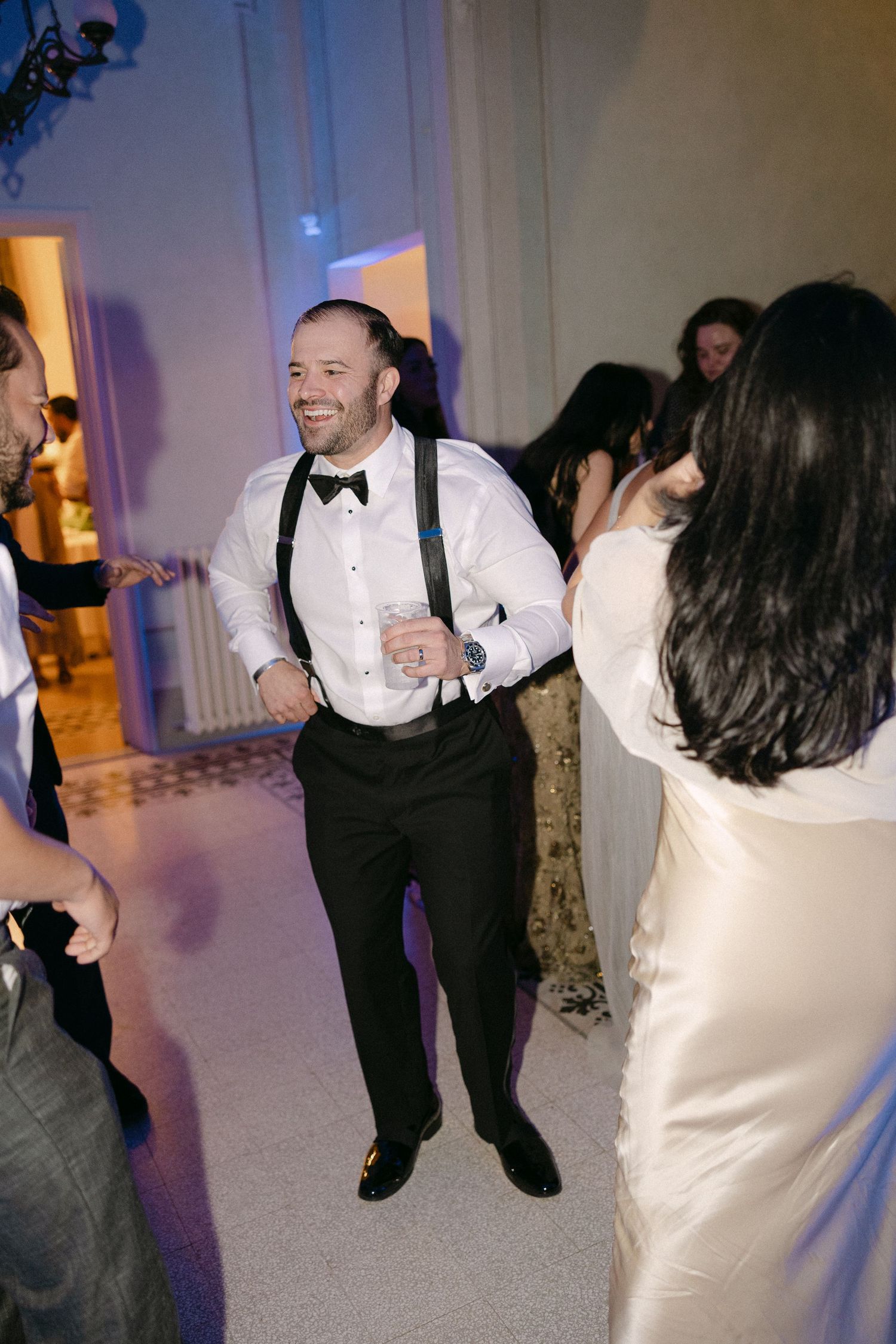 Groom in white shirt with suspenders and bow tie dances joyfully with guests at wedding reception.