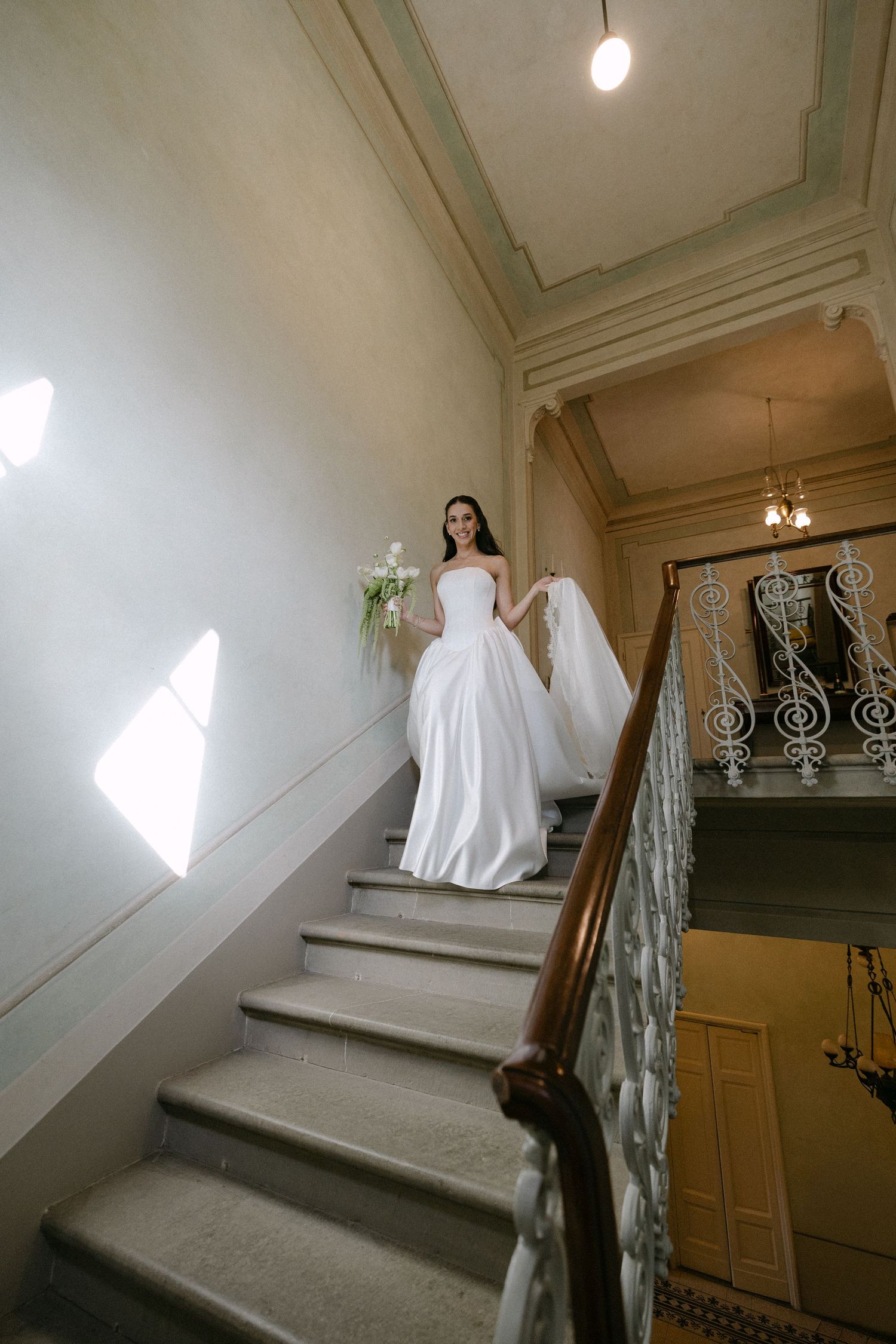 Elegant bride in white gown descending grand staircase with ornate railing in a classic venue.