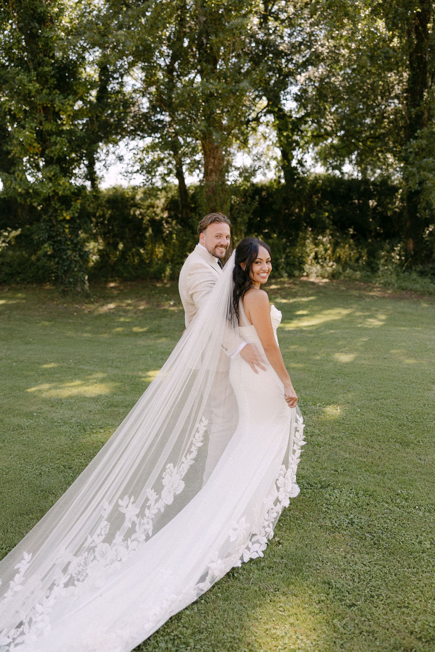 Bride and groom walk on lush lawn, showcasing long lace cathedral veil in golden light.