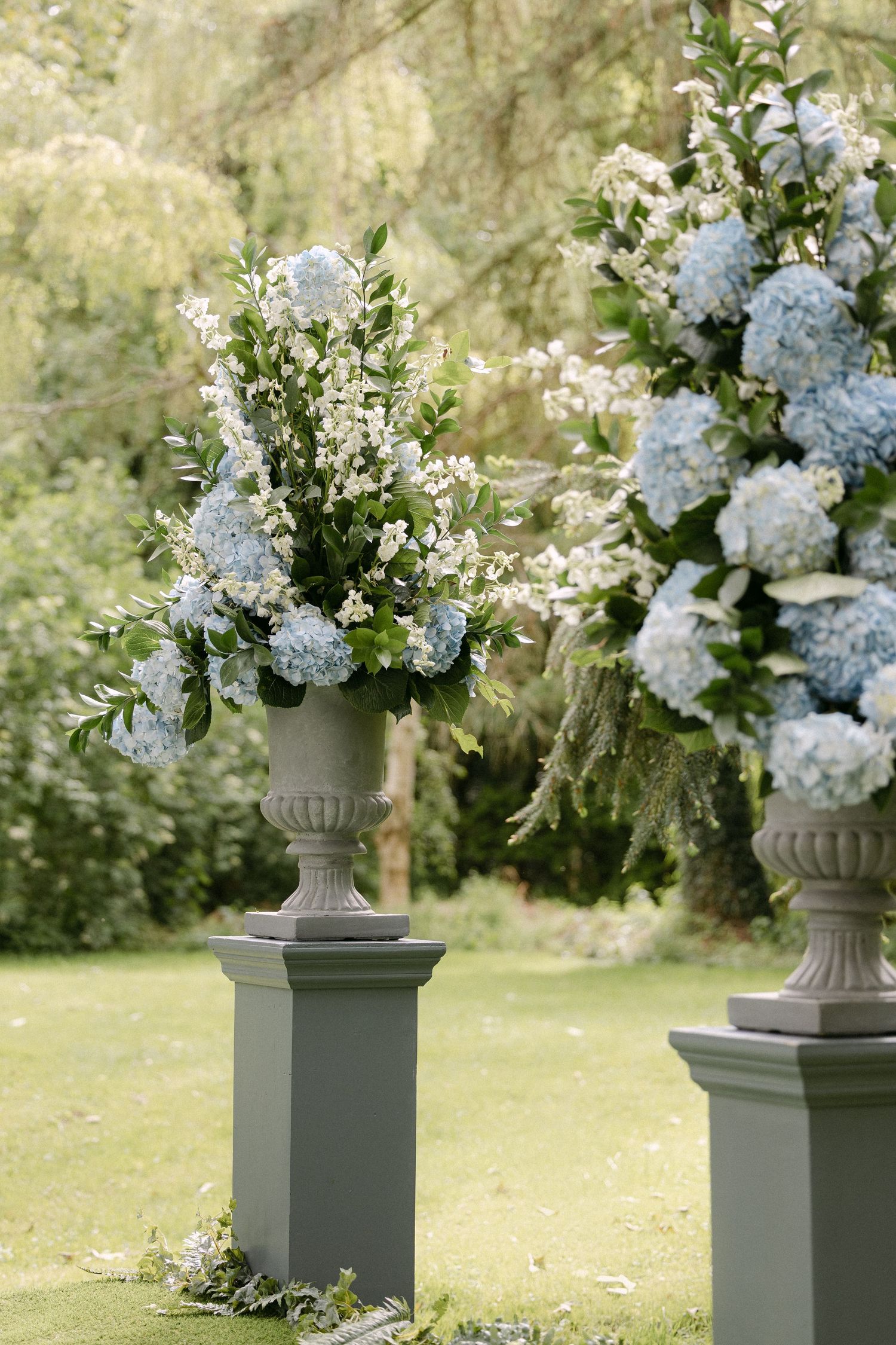 Elegant stone urns with blue hydrangeas and white flowers on grey pedestals in a lush garden wedding ceremony.
