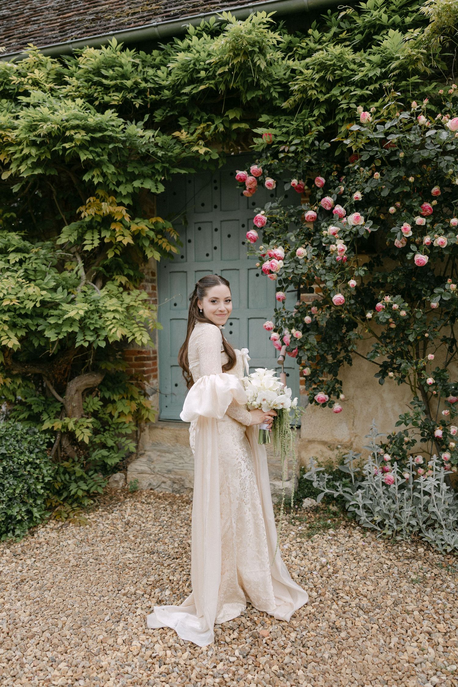 Bride in elegant champagne gown with puff sleeves holds bouquet before a teal door adorned with climbing pink roses.