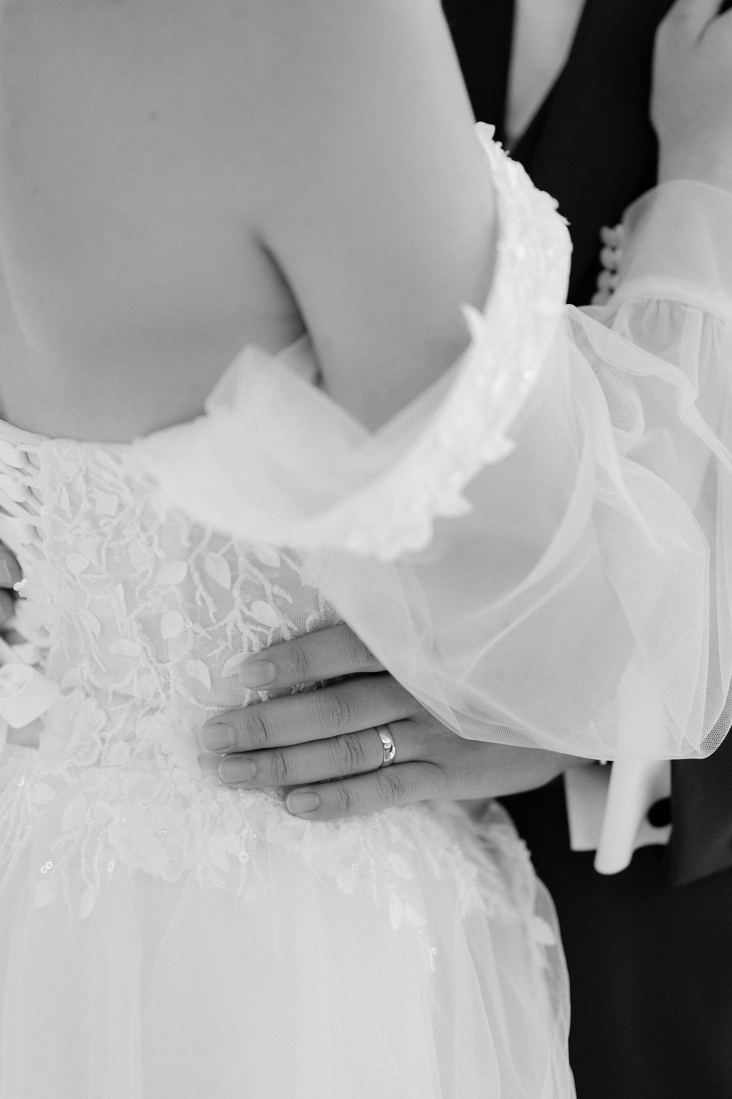 Black and white close-up of bride wearing wedding ring with groom embracing her in elegant lace gown.