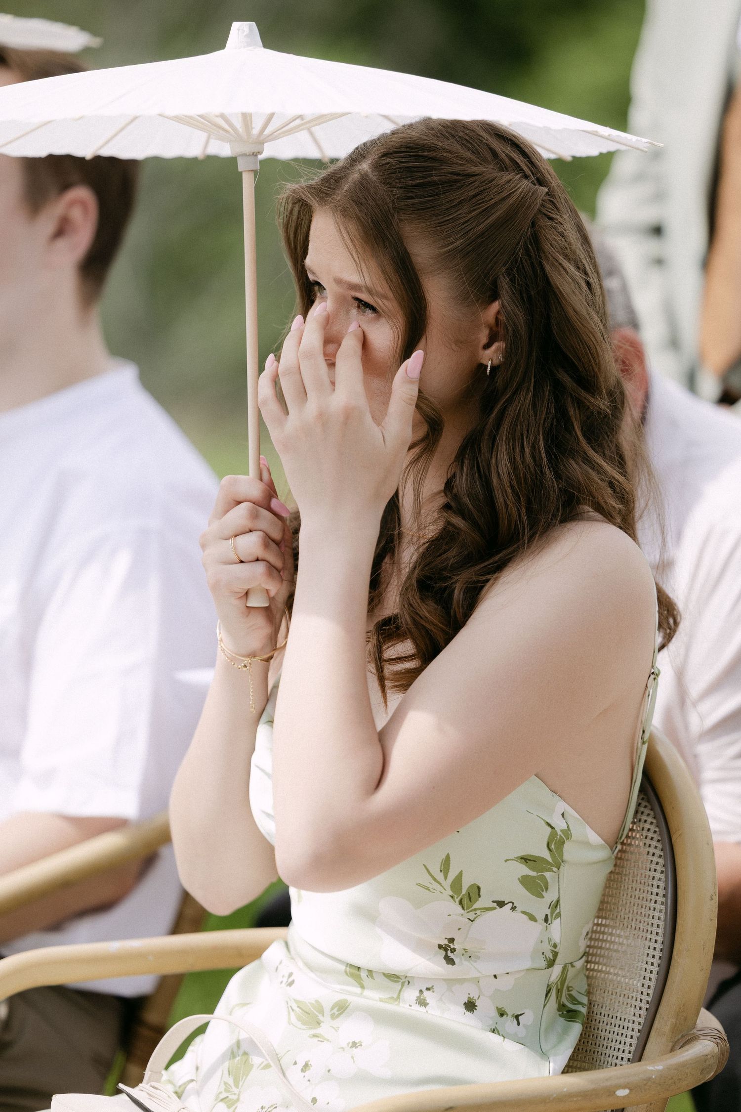Emotional wedding guest in floral dress holds white parasol while wiping tears at outdoor ceremony.