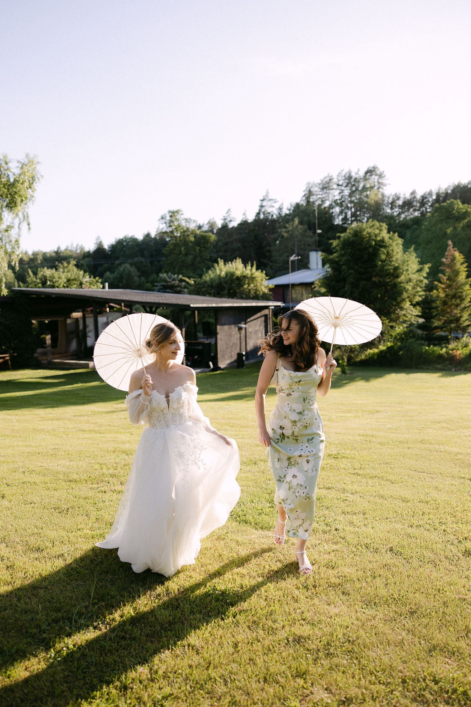 Bride in white gown and bridesmaid in floral dress walk on green lawn holding white parasols.