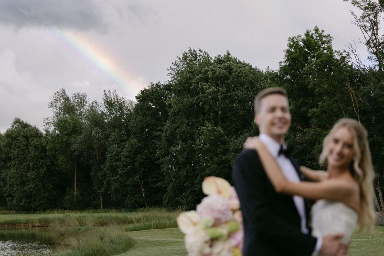 Bride and groom embrace outdoors with a stunning rainbow arching over lush green trees in the background.