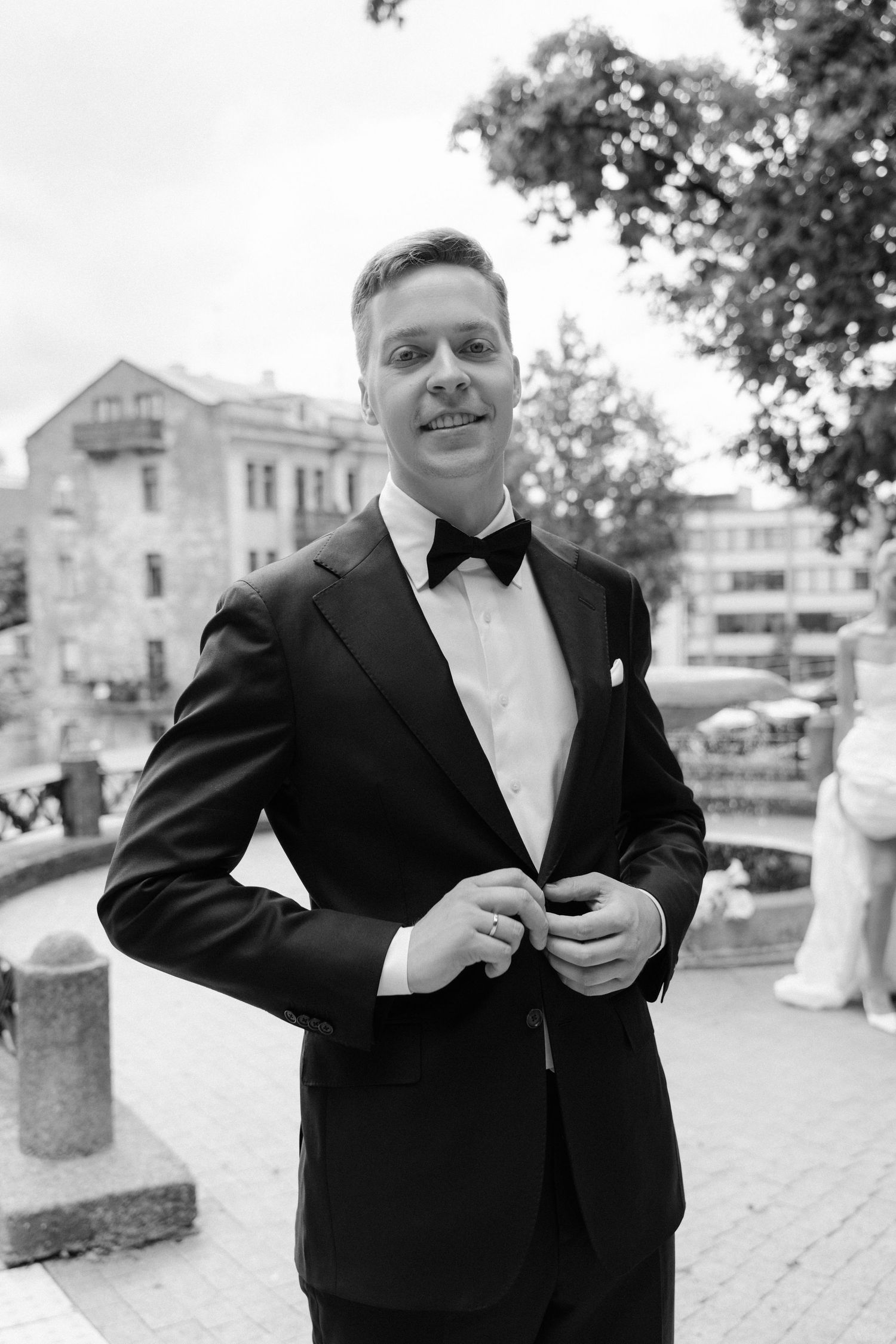 Dapper groom in black tuxedo with bow tie buttoning jacket outdoors in black and white photo.