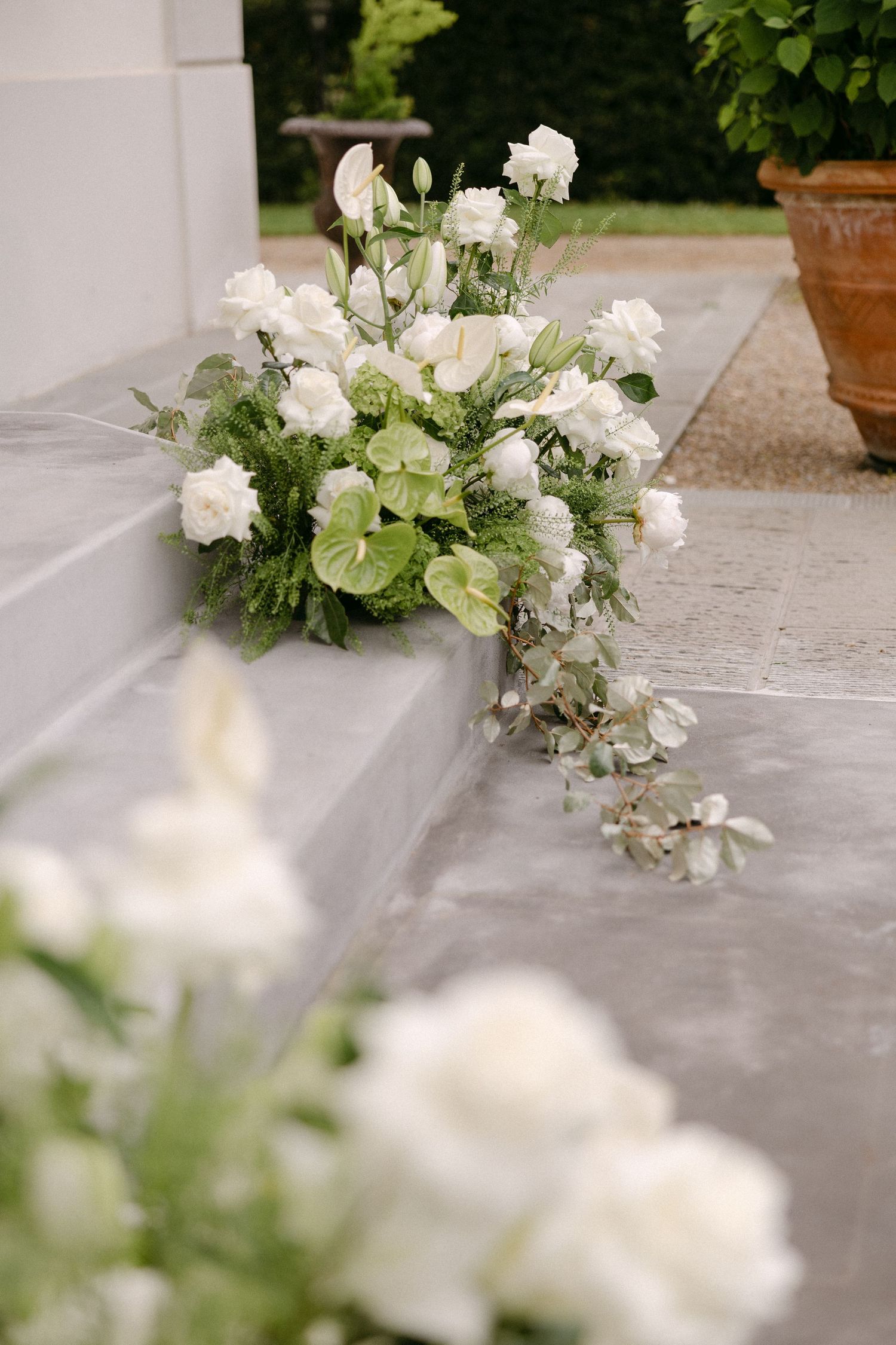 Lush white wedding floral arrangement with roses, anthurium, and greenery cascading down stone chapel steps outdoors.