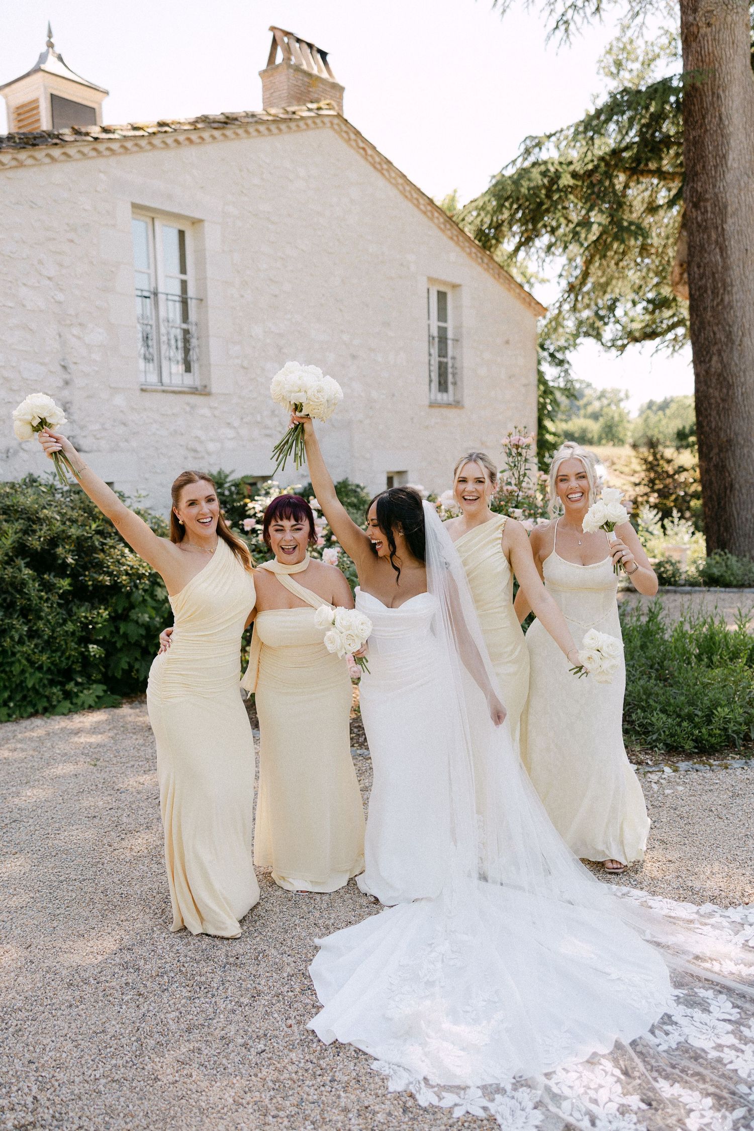 Four bridesmaids in cream dresses celebrate joyfully with bride outside a French stone venue.
