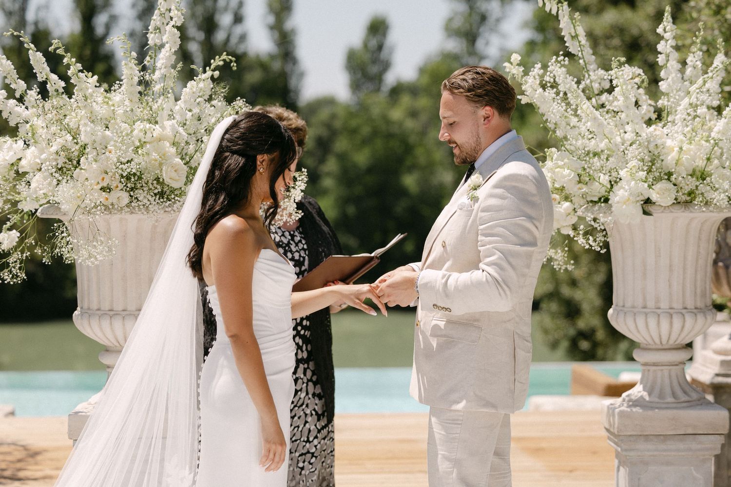 Groom placing wedding ring on bride's finger during outdoor ceremony with white floral urns and pool backdrop.