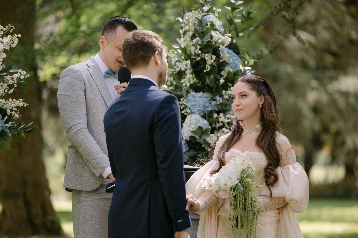 Bride in blush off-shoulder gown holding white bouquet during outdoor wedding ceremony in a park.