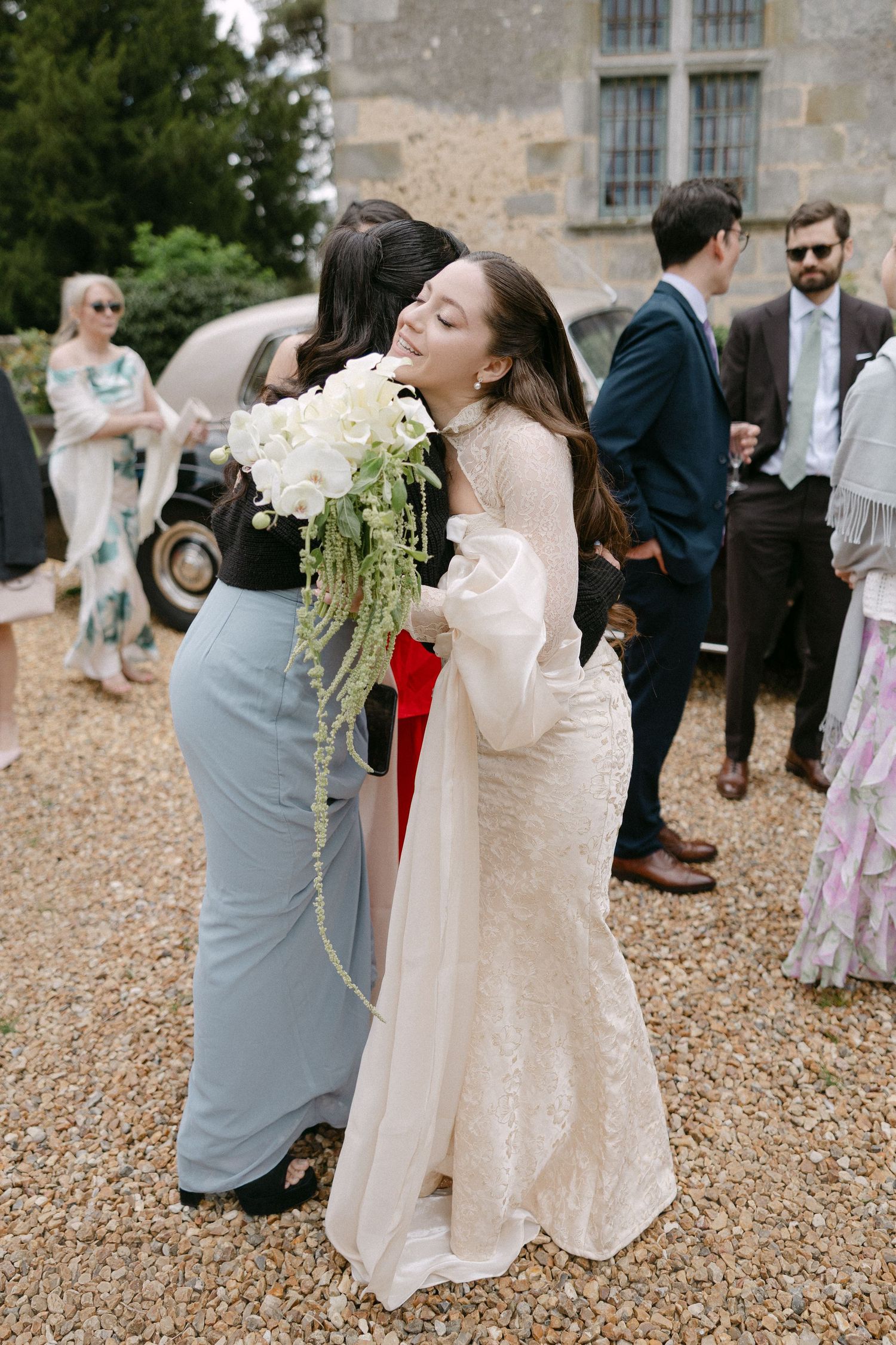 Bride in lace gown hugs a guest while holding white floral bouquet at wedding reception.