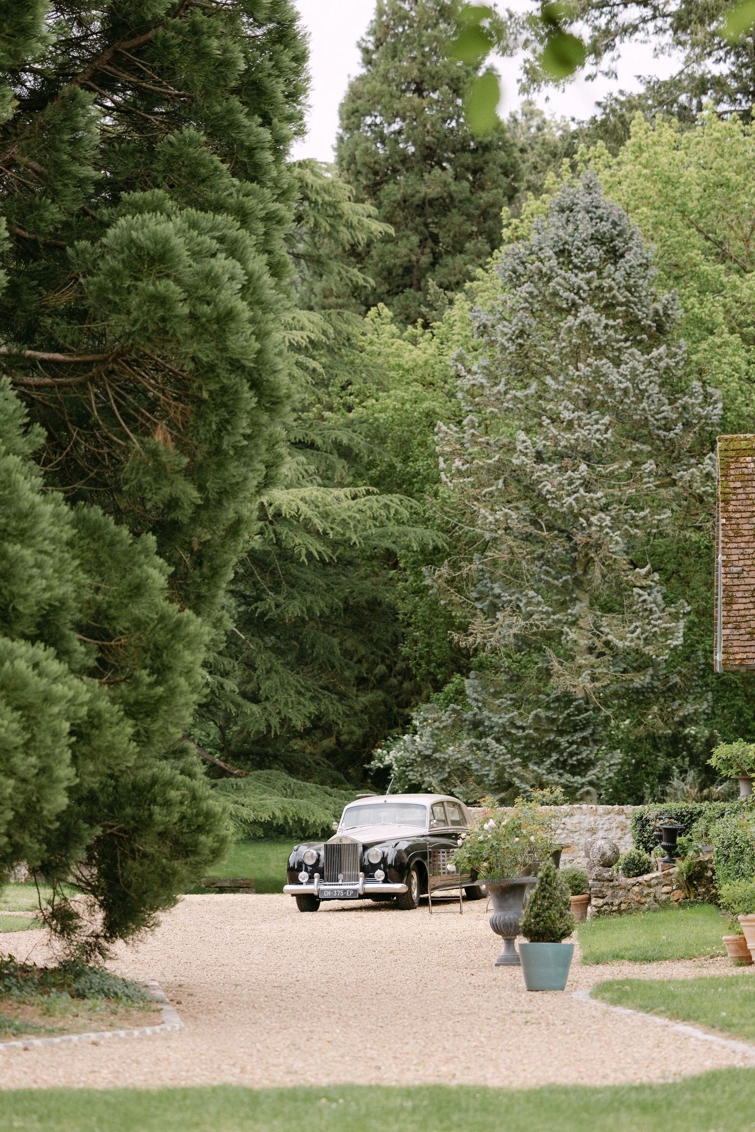Vintage black classic car parked on gravel driveway surrounded by lush green trees at estate.