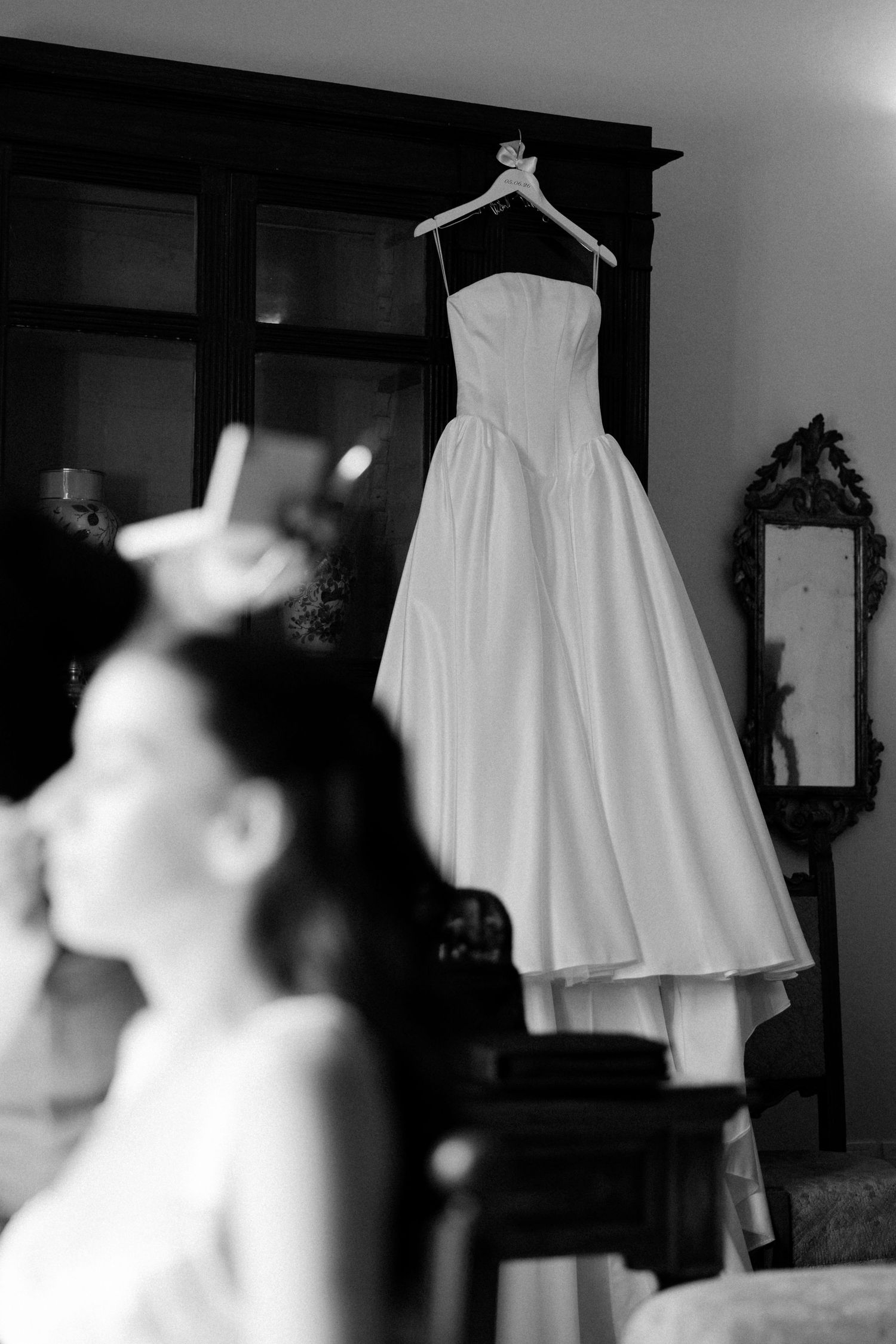 Black and white photo of a wedding dress hanging on a hanger while a bride gets her makeup done in the foreground.