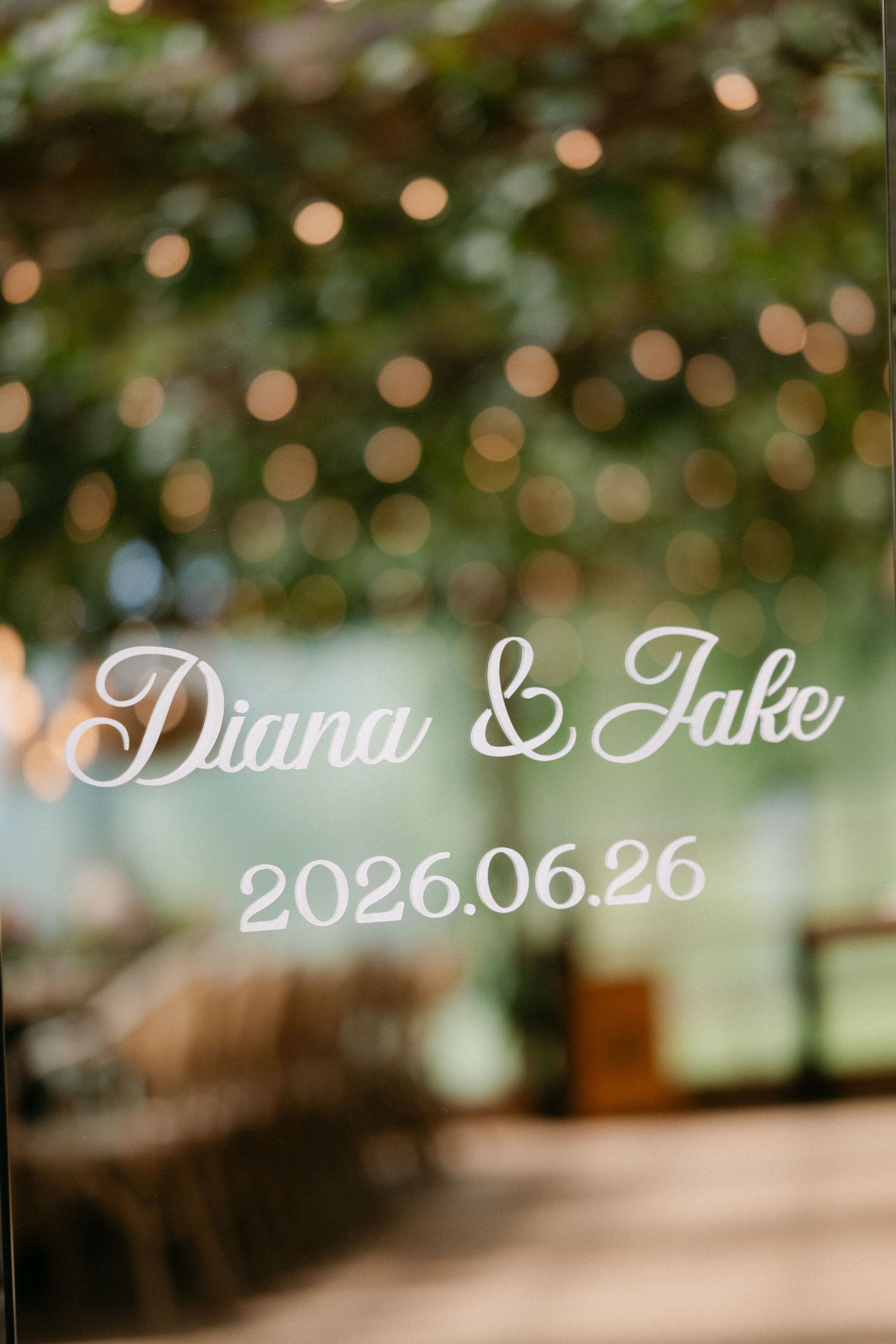 Elegant wedding sign with cursive text reading Diana & Jake 2026.06.26 against a bokeh greenery background.