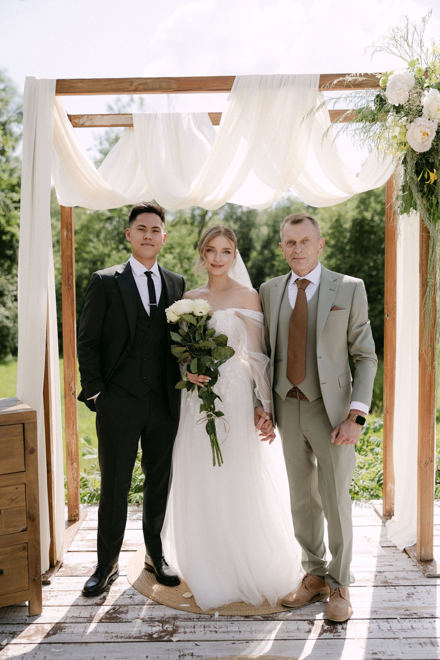 Bride holding white roses poses with groom and officiant under wooden wedding arch.