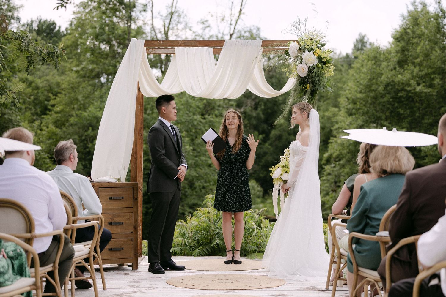 Outdoor wedding ceremony with bride, groom, and officiant standing under a wooden arch draped with white fabric and floral arrangements.