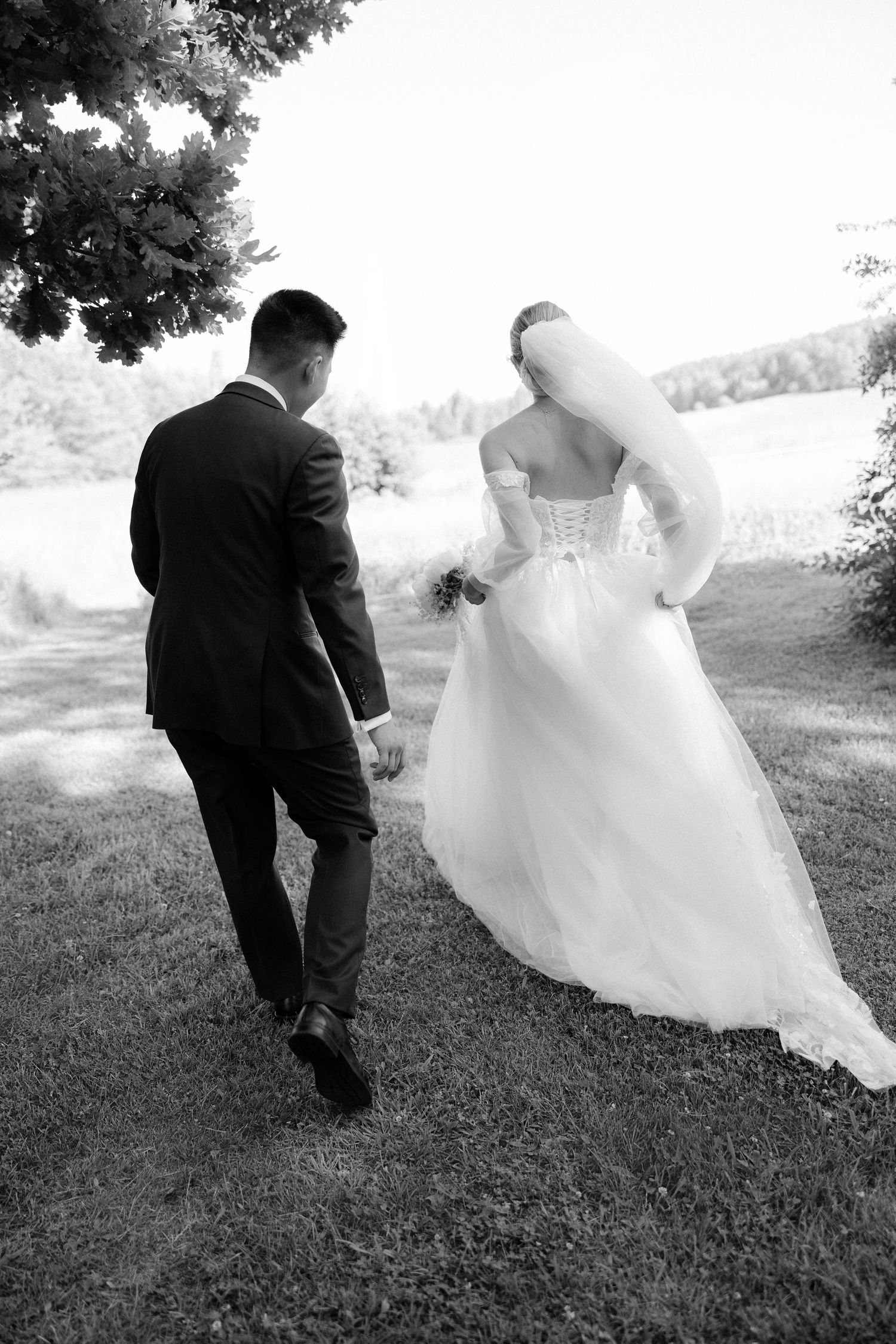 Black and white photo of bride and groom walking hand in hand across green lawn outdoors.