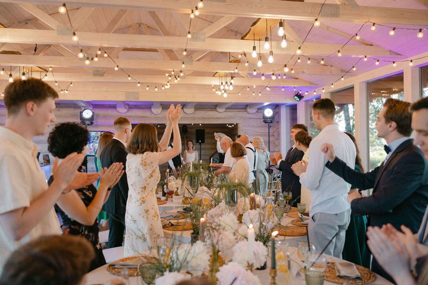 Newlyweds enter wedding reception as guests applaud beneath string lights in elegant decorated venue.
