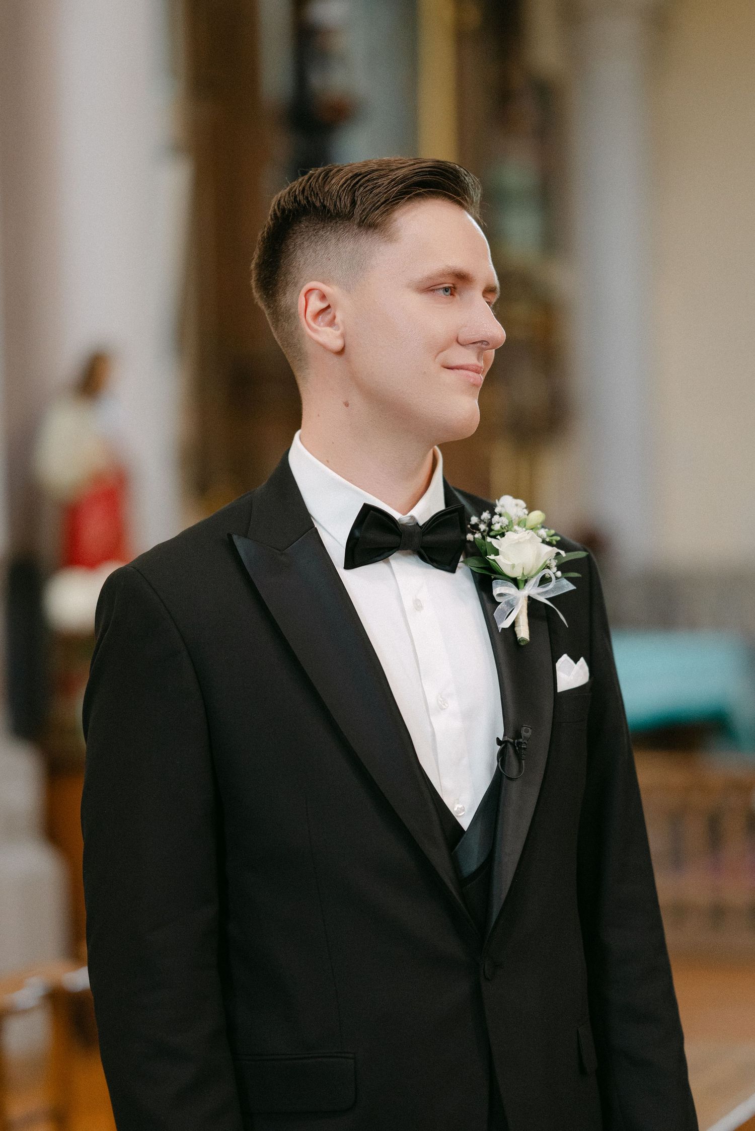 Handsome groom in black tuxedo with white boutonniere smiling inside elegant church wedding venue.
