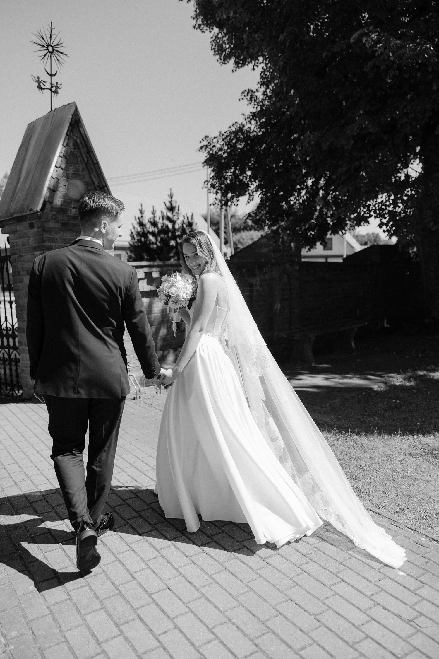 Black and white photo of bride and groom walking hand in hand on cobblestone path.