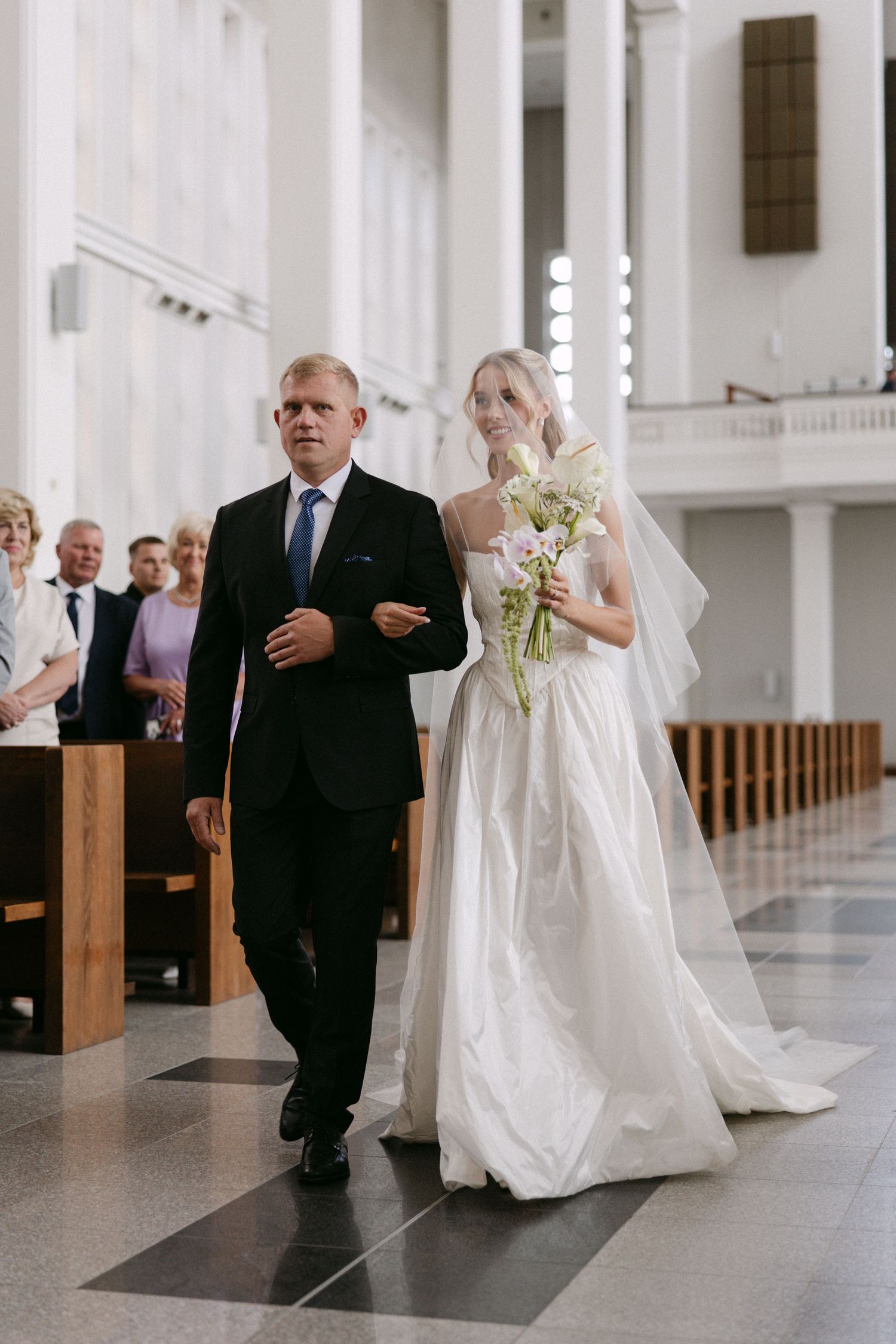 Bride in white gown and veil walks down church aisle with escort during wedding ceremony.