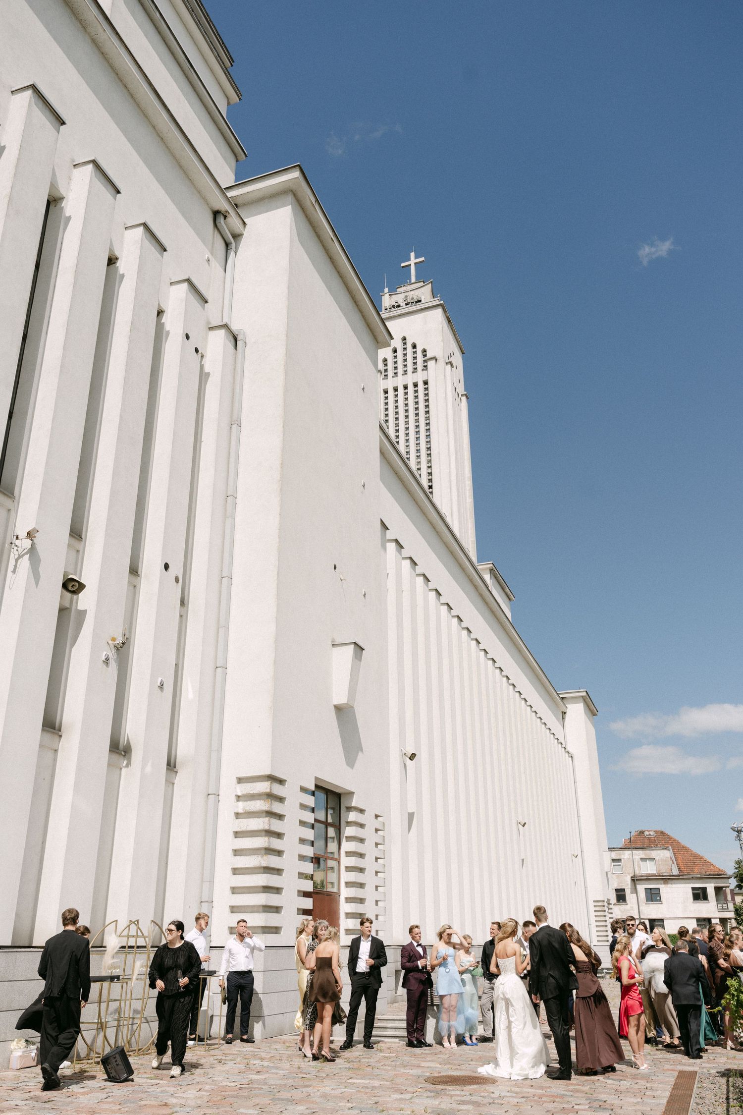 Wedding guests mingling outside a white modernist church with a cross on the tower under a clear blue sky.