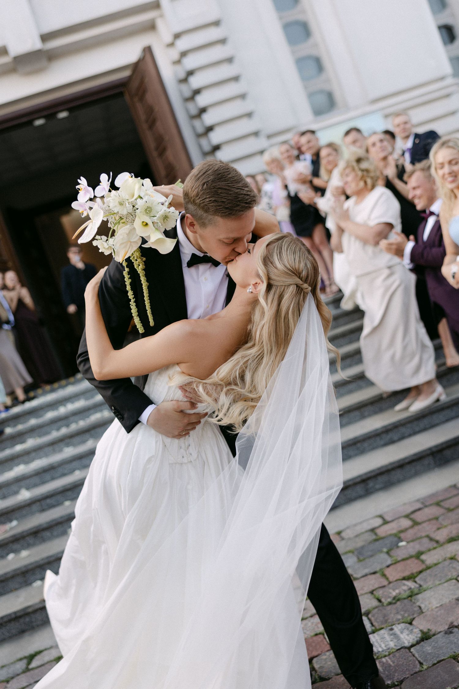 Bride and groom share a romantic kiss on church steps holding white bouquet while wedding guests celebrate behind them.