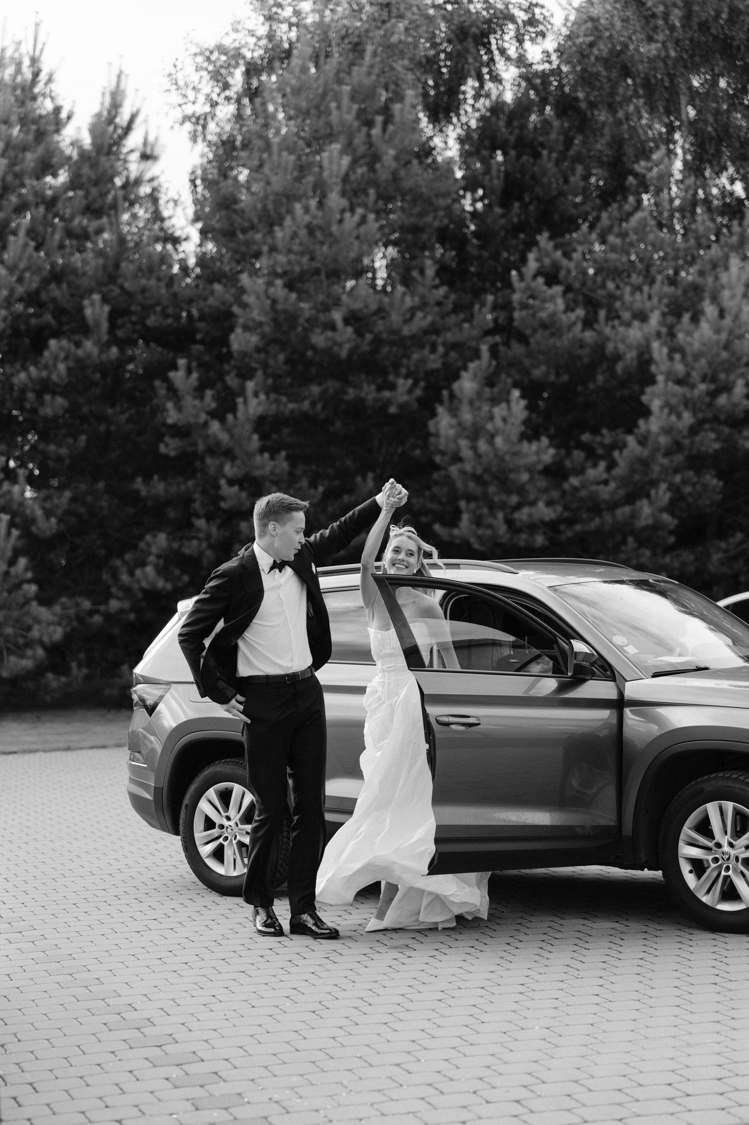 Black and white photo of bride and groom dancing beside a luxury SUV with evergreen trees in background.