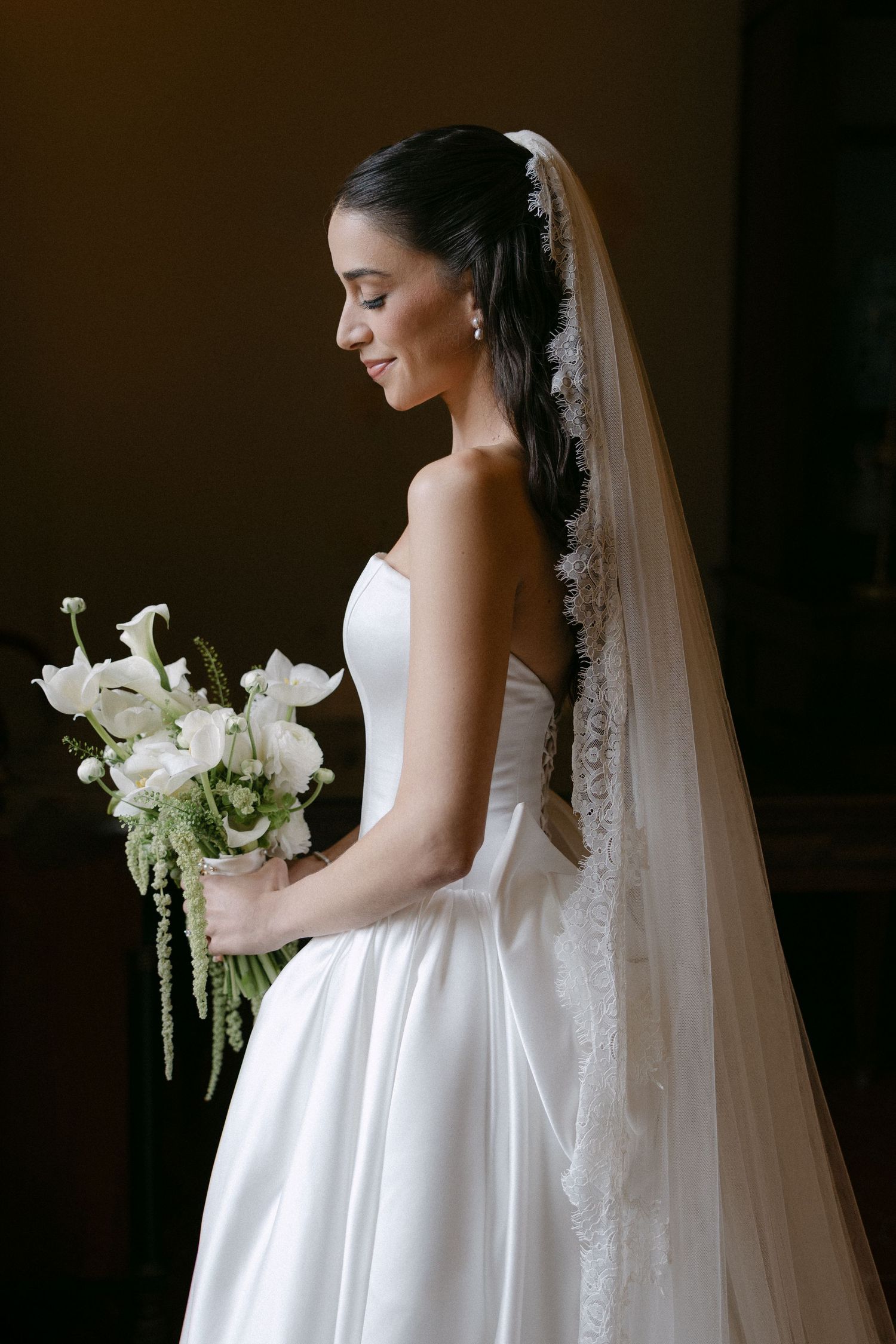 Elegant bride in strapless white gown holding white calla lily bouquet with long lace-trimmed veil, smiling softly.