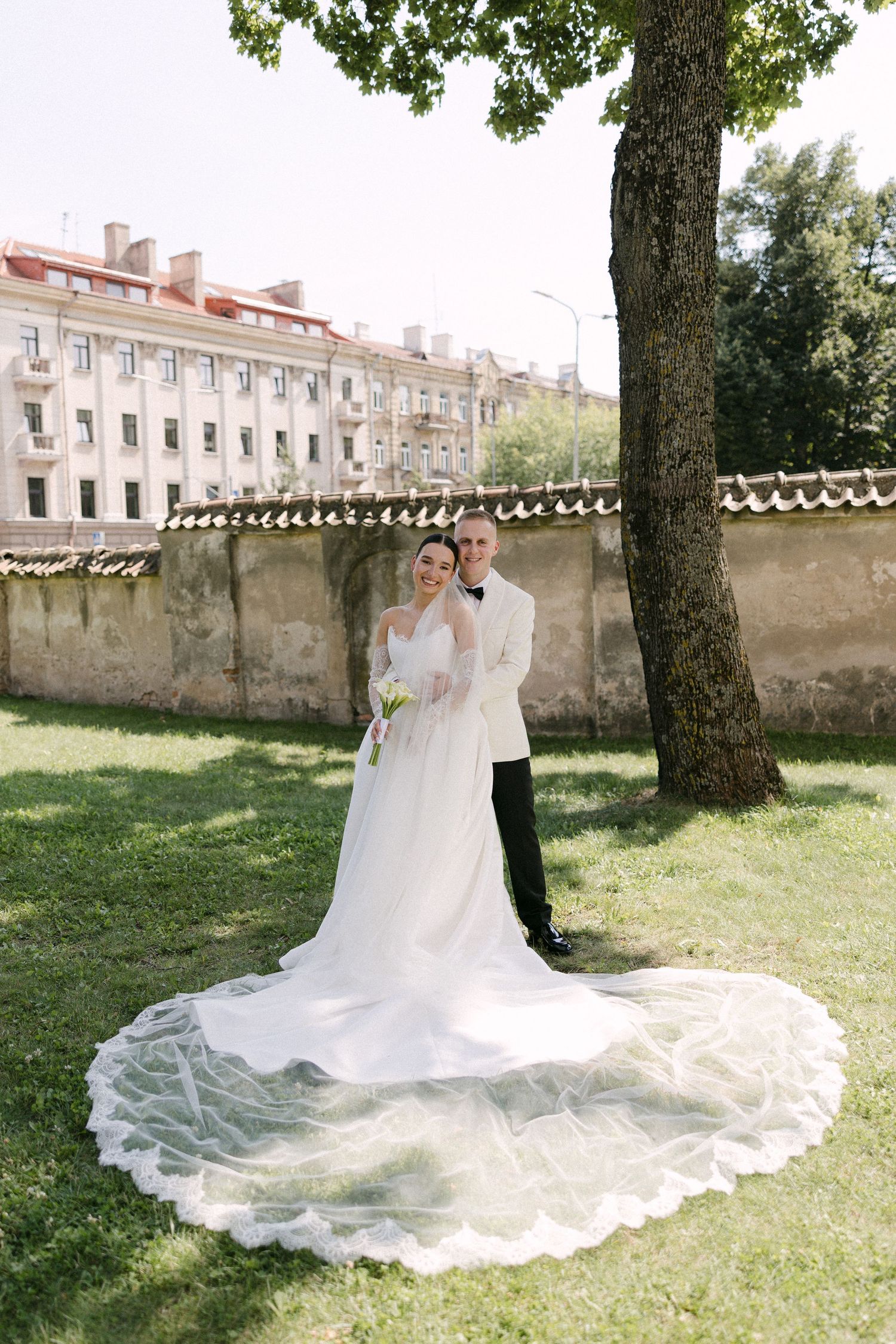 Wedding couple posing under a large tree on green lawn with historic European buildings in background.