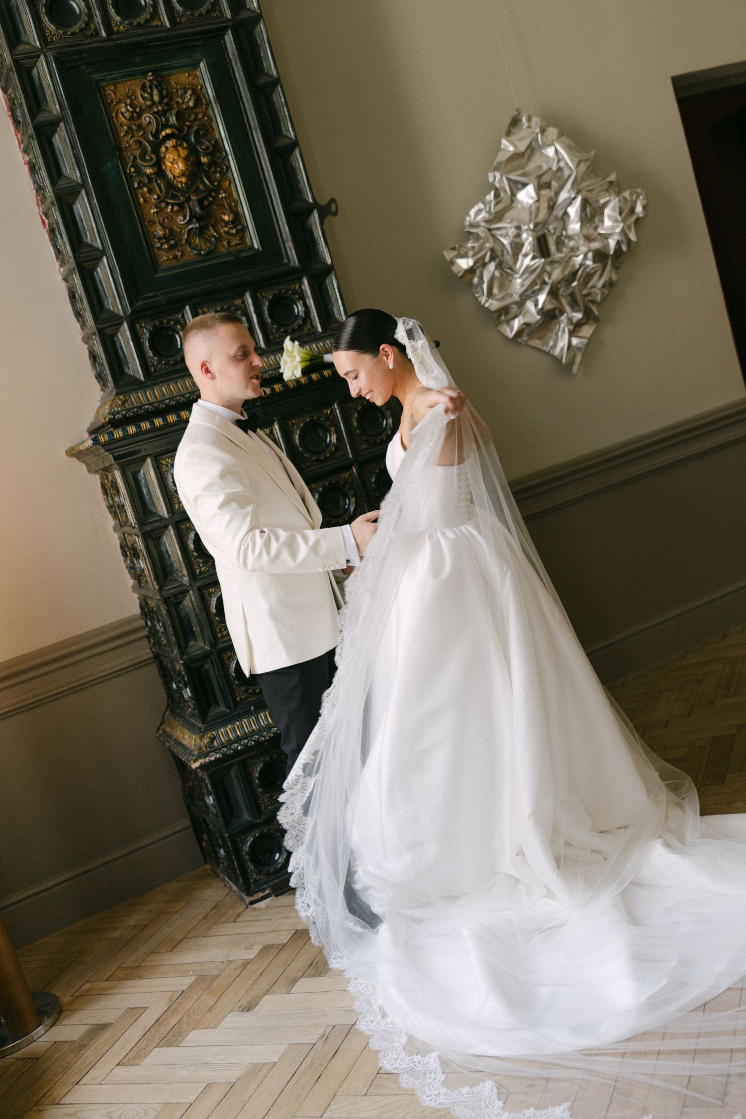 Groom in white jacket holds bride's hand tenderly near ornate fireplace in elegant interior.