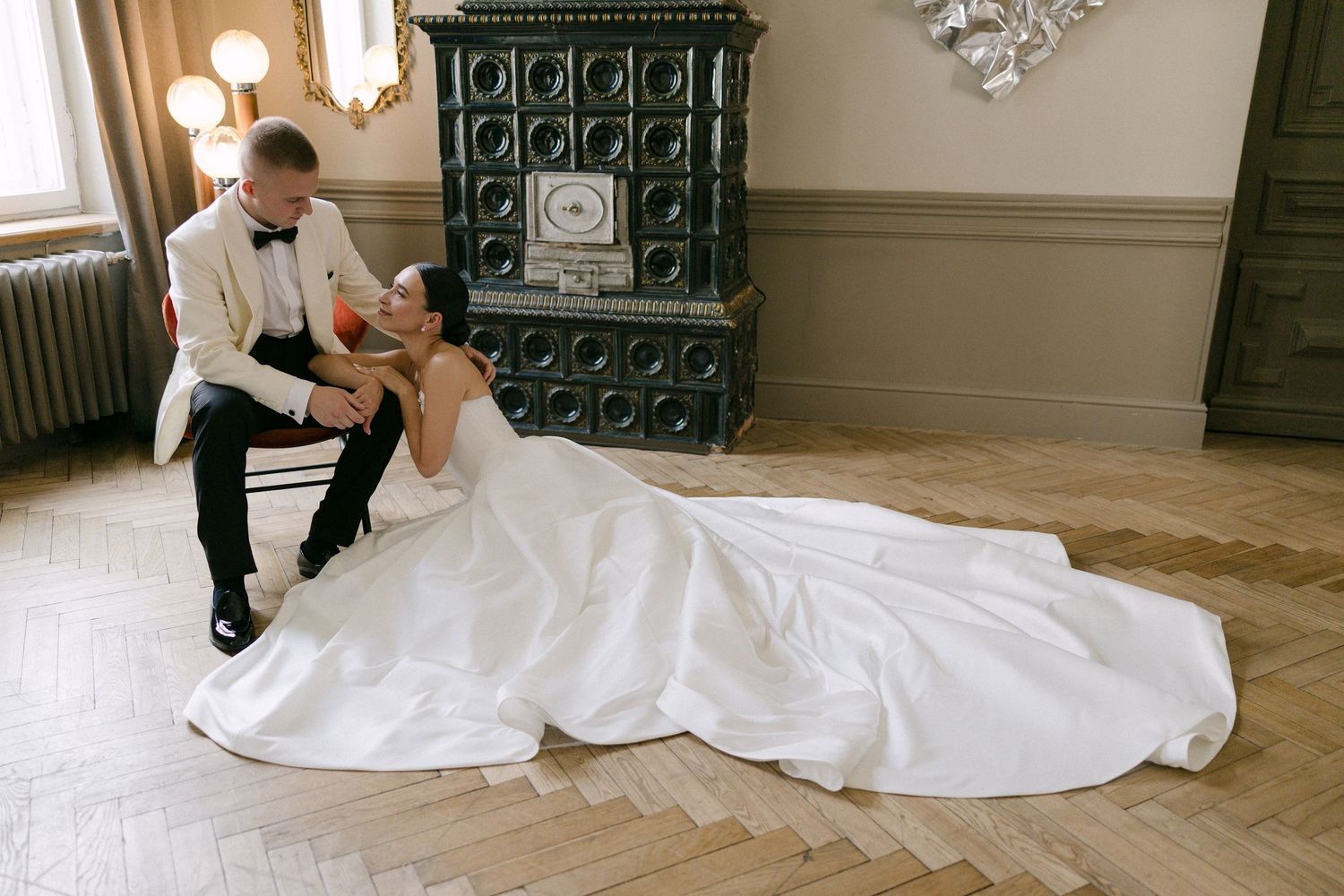 Bride in flowing white gown reclines on parquet floor gazing up at groom in white tuxedo jacket.