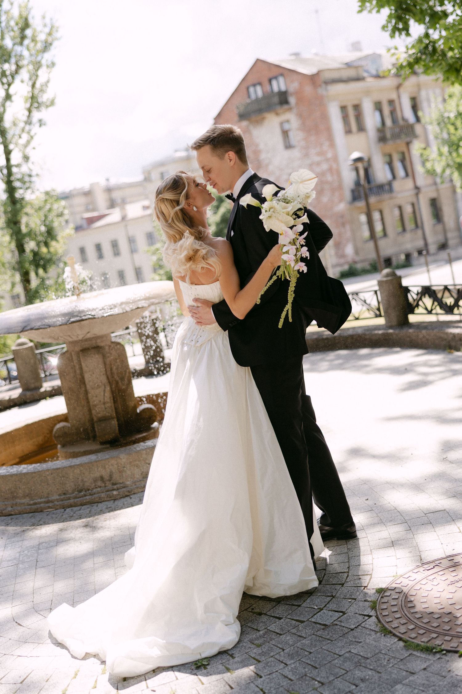Bride and groom sharing a romantic kiss near a stone fountain in a sunlit European city square.