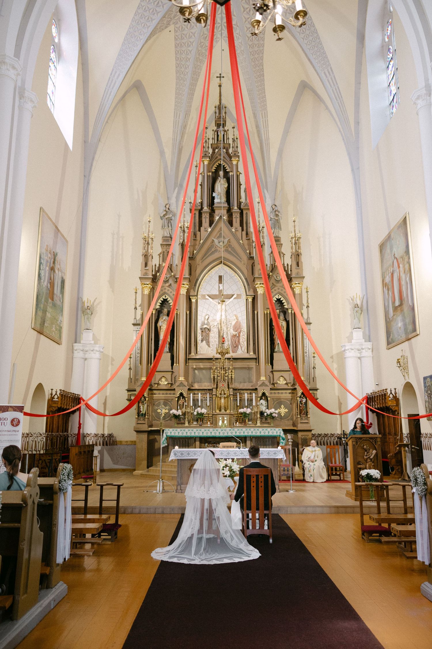 Bride and groom kneeling at ornate Gothic church altar decorated with red ribbons during ceremony.