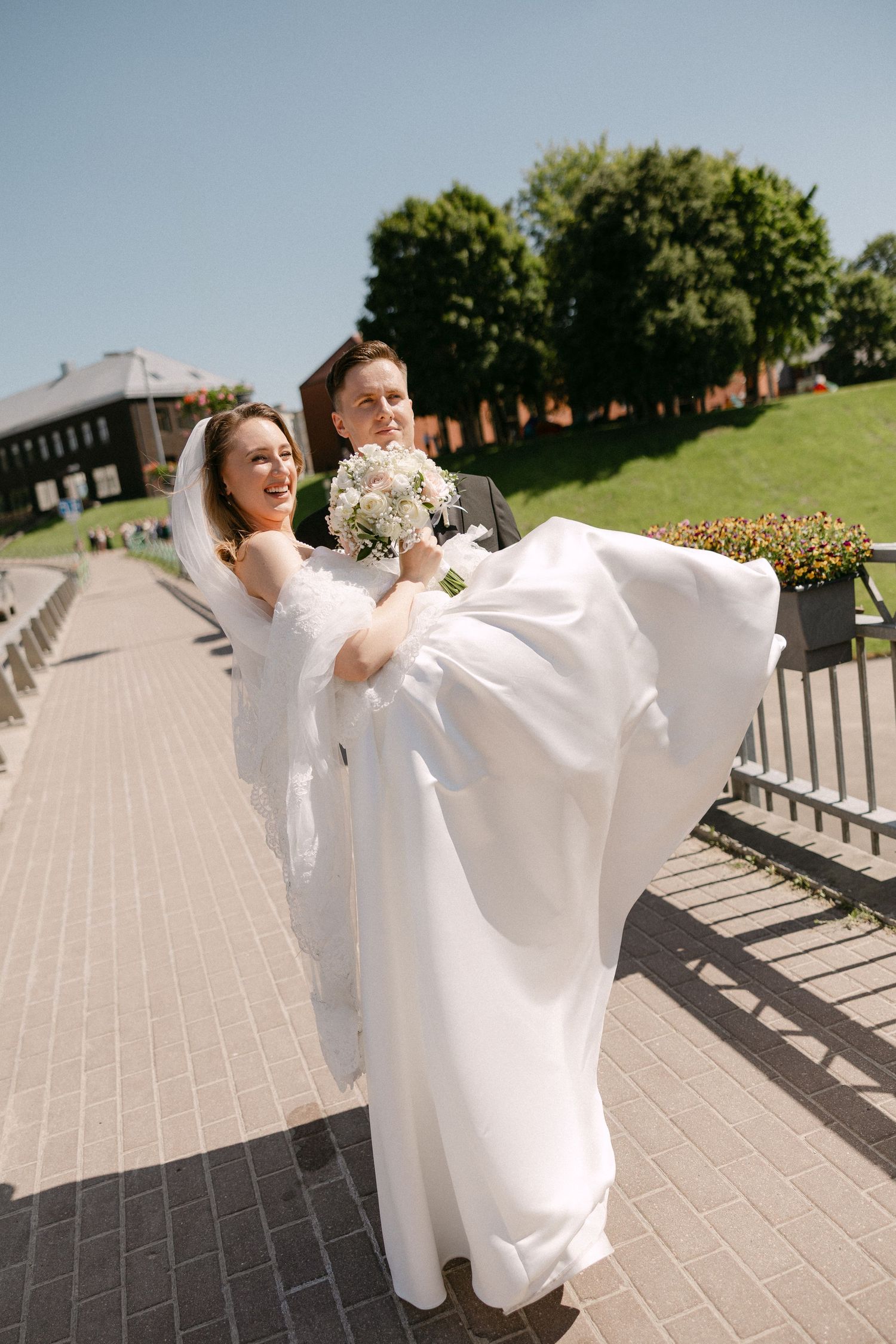 Groom lifts smiling bride in white wedding dress and veil on sunny outdoor walkway with green trees.