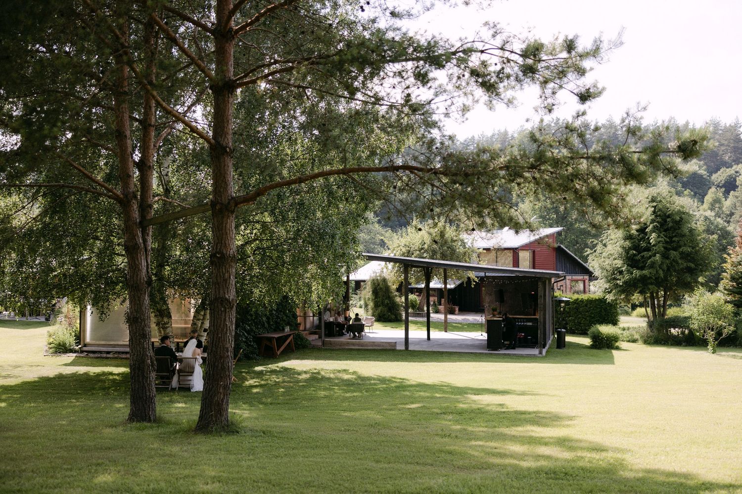 Scenic outdoor wedding venue with tall pine trees, lush green lawn, and a covered pavilion in the background.