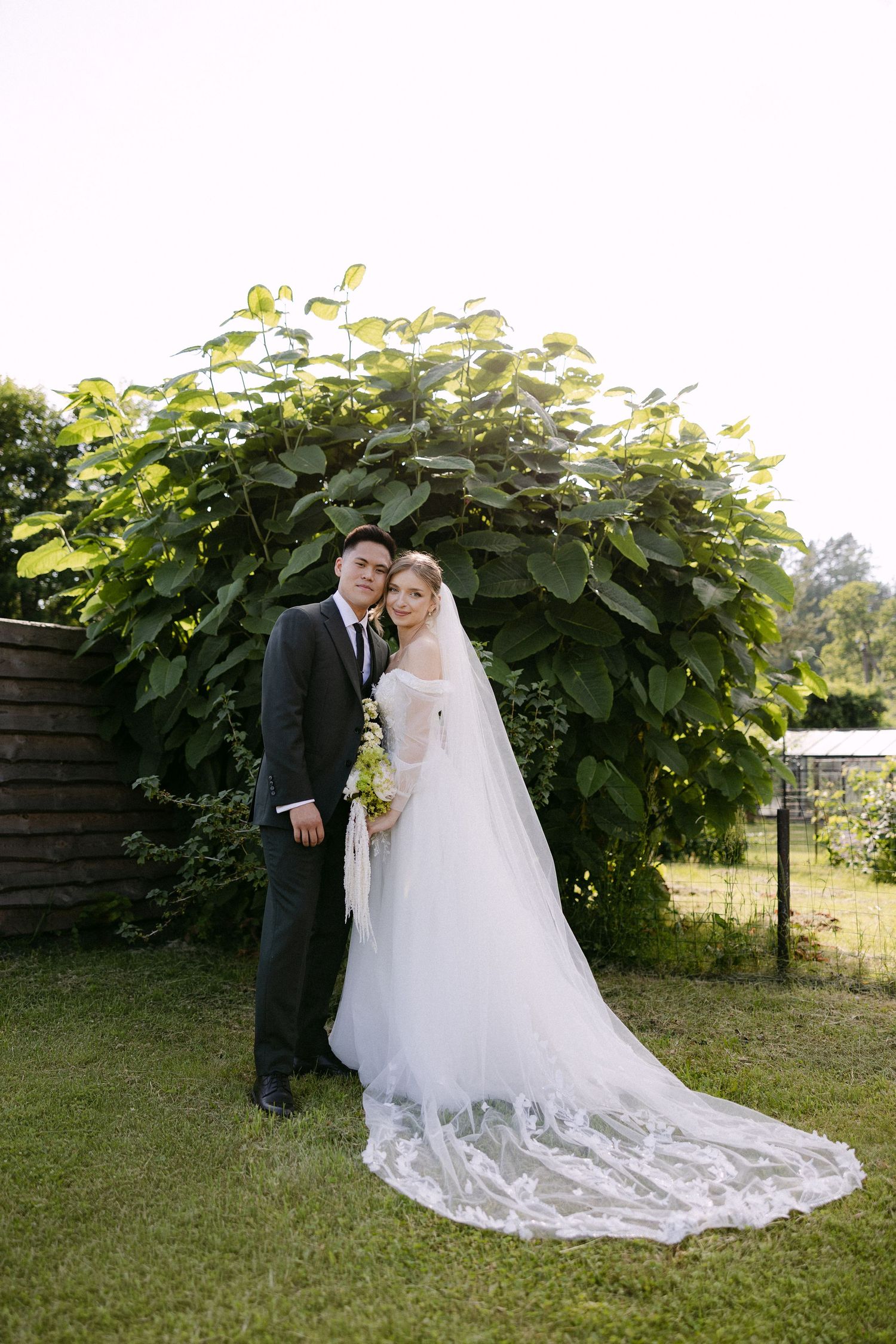 Bride and groom pose together in front of lush green foliage arch at outdoor garden wedding.