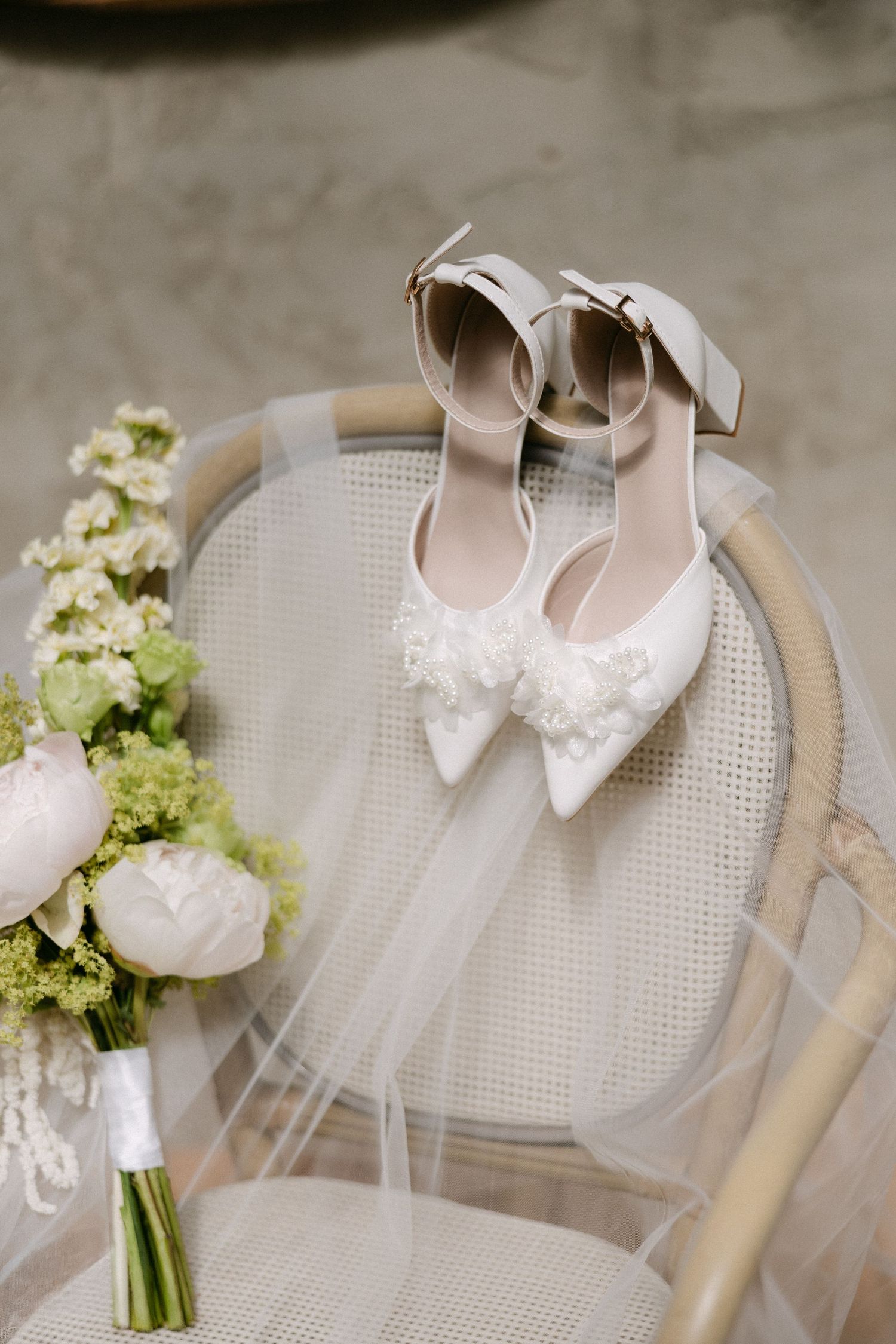 White bridal heels with floral embellishments and ankle straps displayed on a chair with a wedding bouquet.