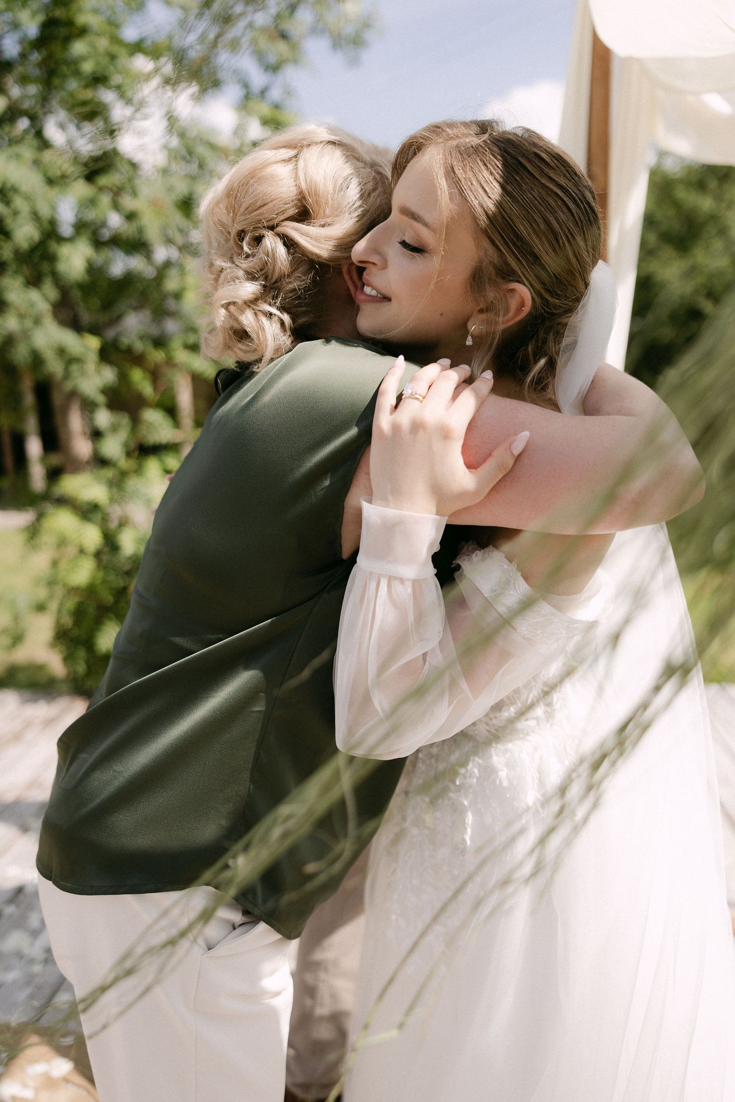 Bride in white gown smiles while embracing guest in green jacket near wooden wedding arch outdoors.