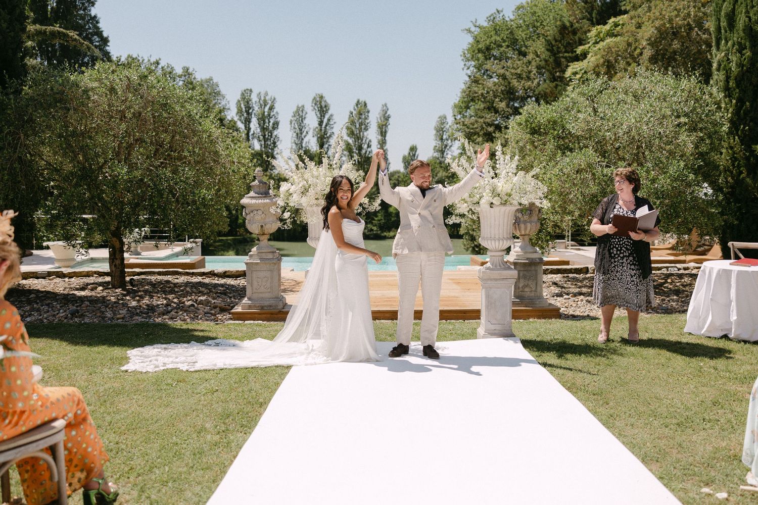 Newlyweds celebrating with raised hands at outdoor wedding ceremony on white aisle with pool and garden backdrop.