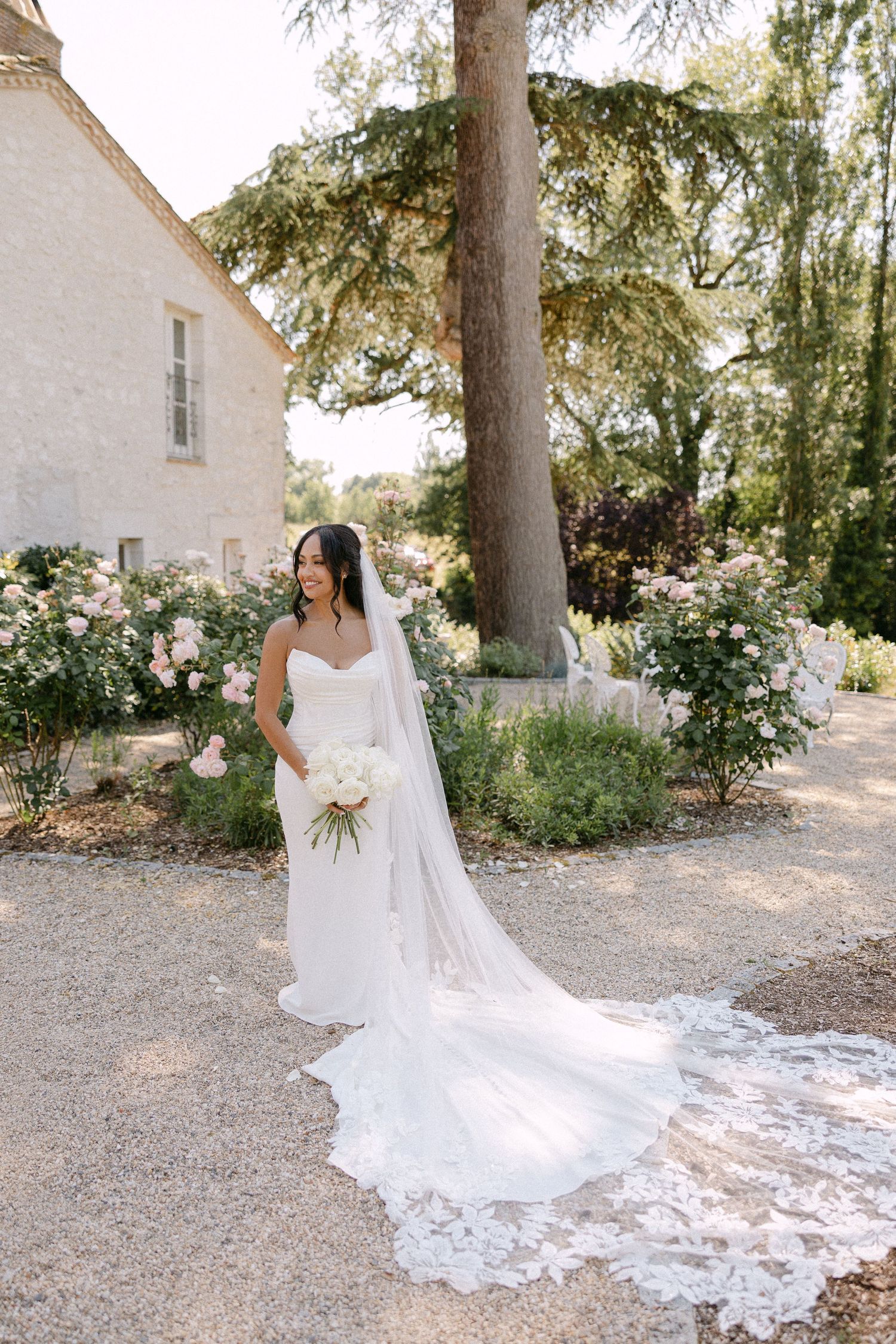 Bride in strapless gown with long lace-trimmed veil poses in chateau garden with roses.
