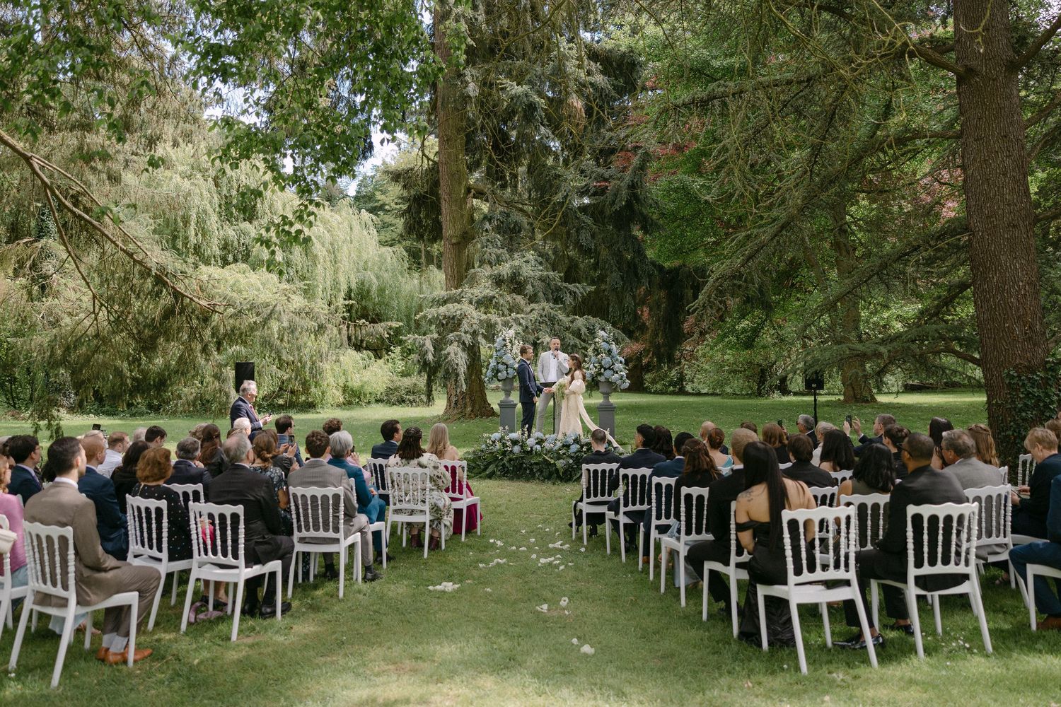 Outdoor wedding ceremony with guests seated in white chairs beneath large trees in a garden.