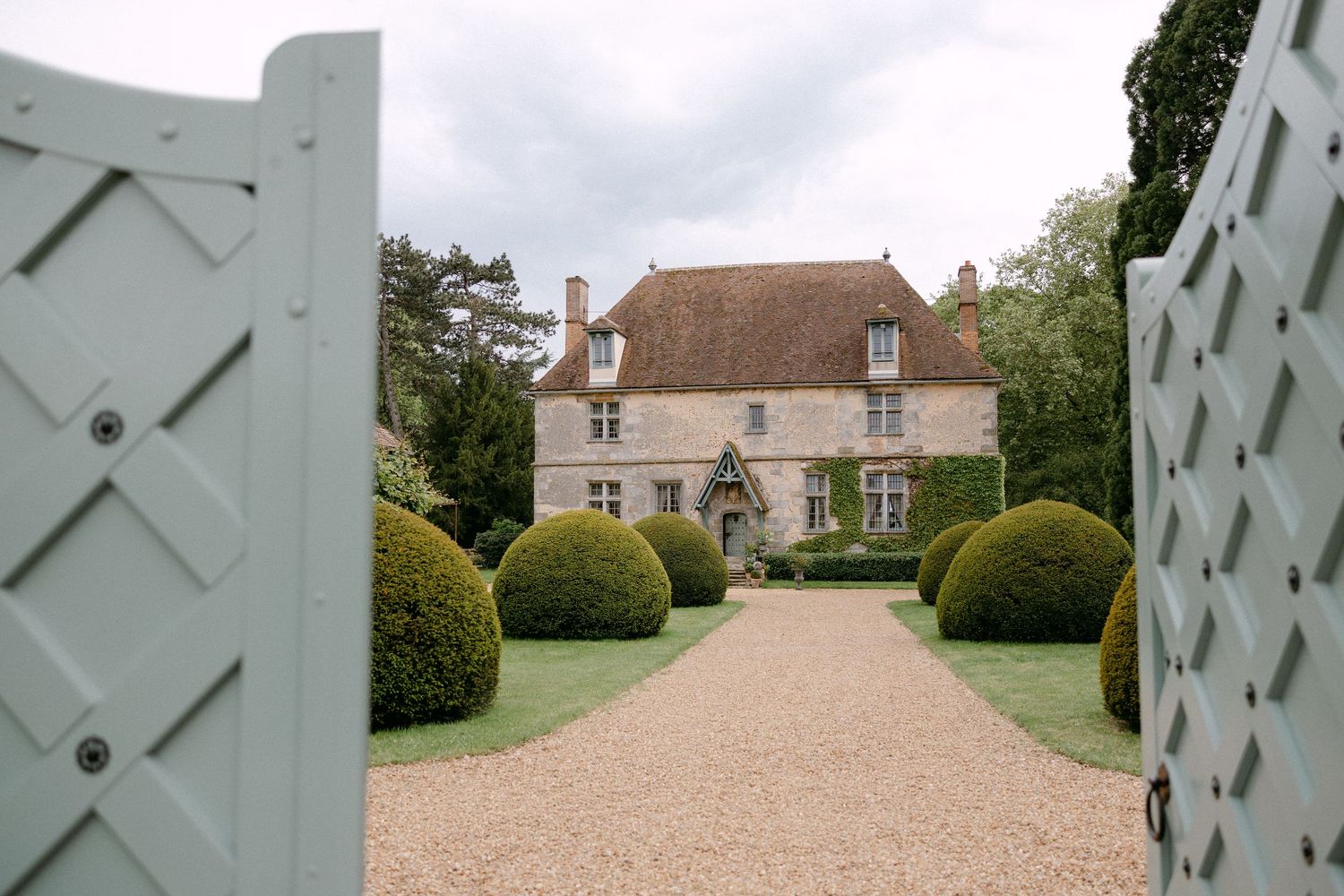 Elegant French manor house viewed through open gates with manicured topiary hedges lining a gravel driveway.
