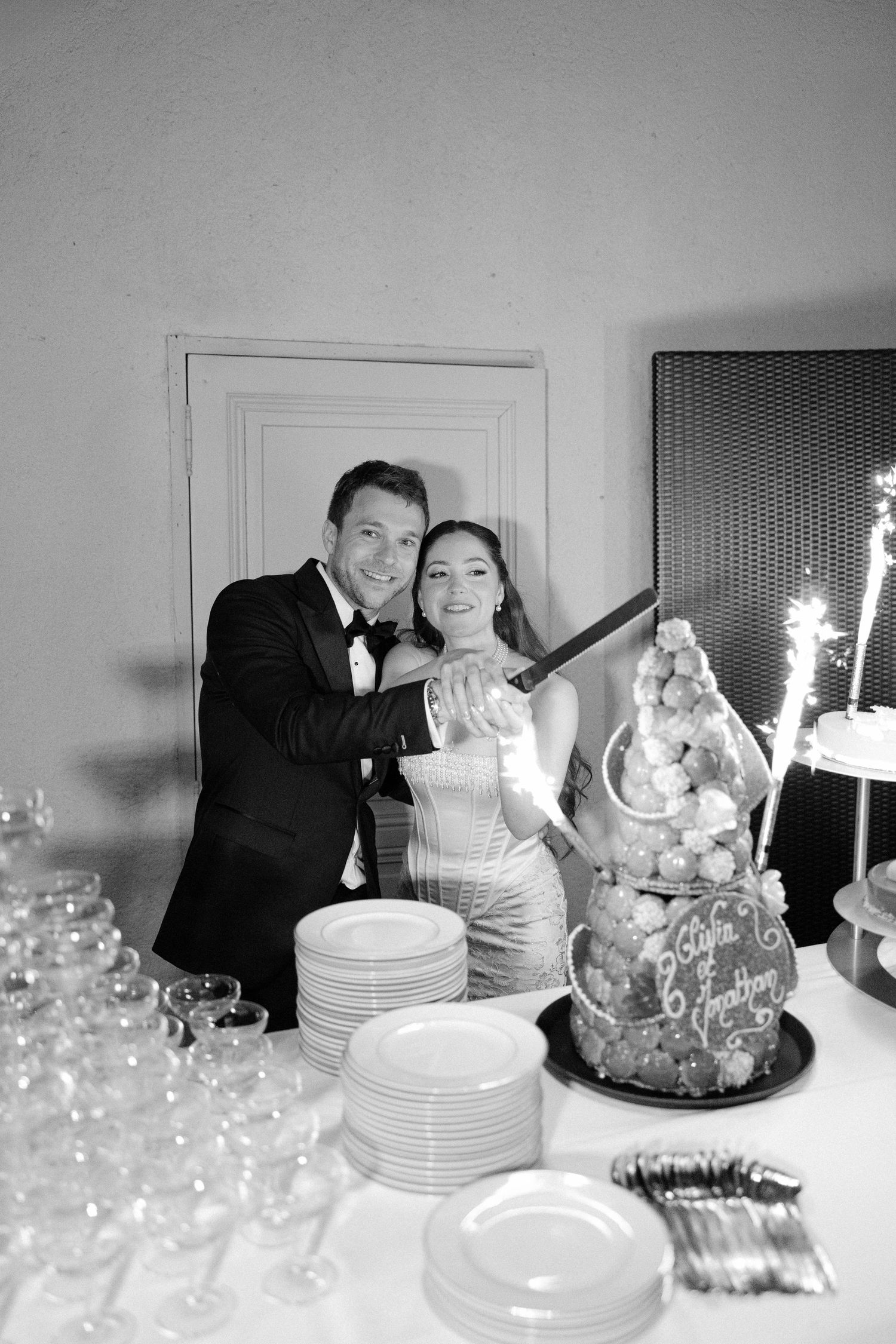 Black and white photo of a couple cutting their wedding cake together at a reception table with glasses and plates.