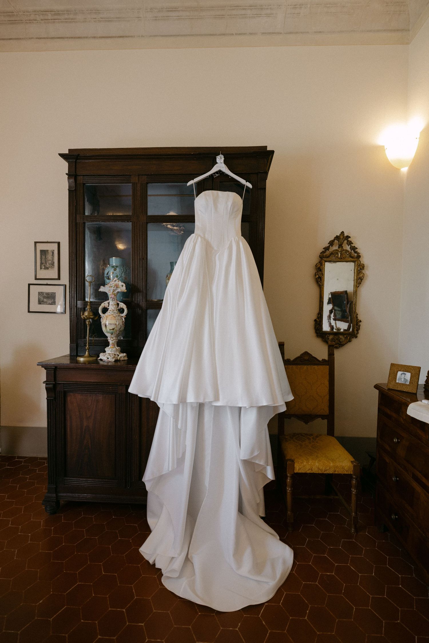 Elegant strapless white ball gown wedding dress hanging on antique wooden armoire in vintage Italian villa room.