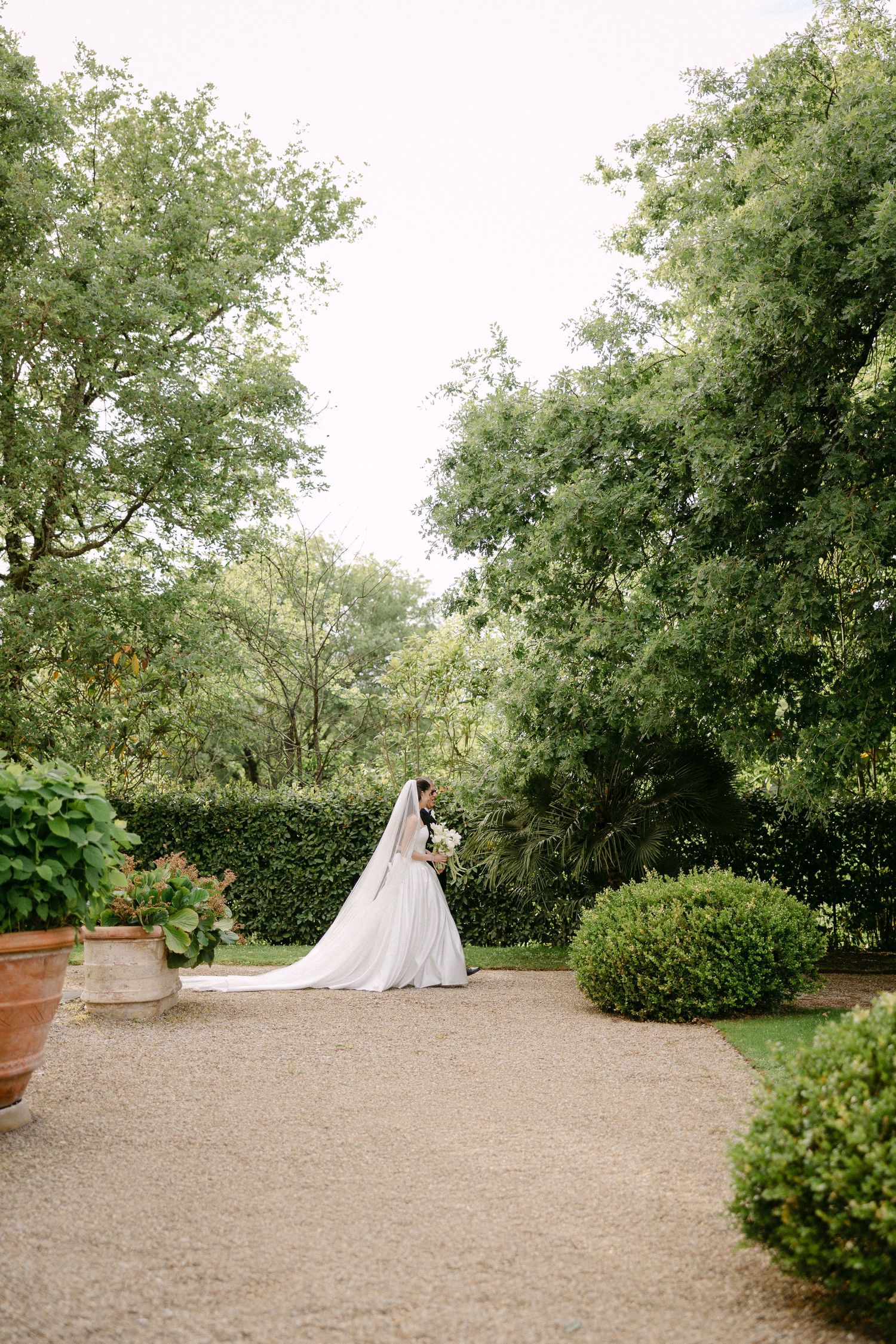 Bride and groom embrace on gravel path surrounded by lush green trees and manicured hedges at garden wedding.