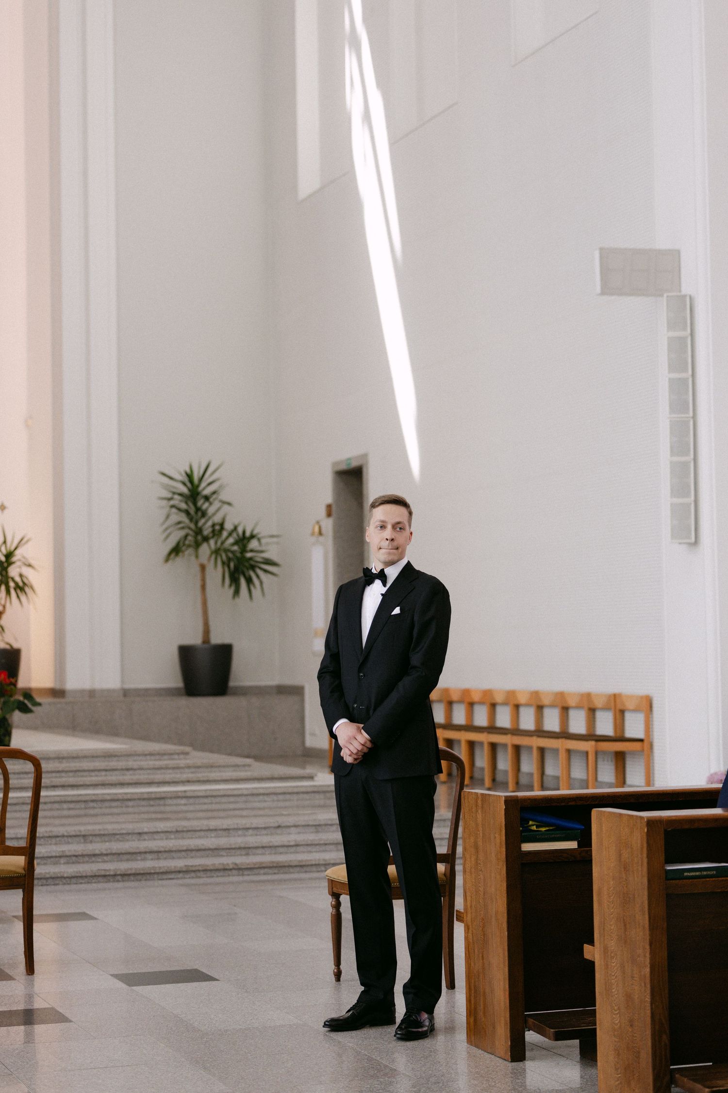 A groom in a black tuxedo stands smiling in a bright modern church interior before his wedding ceremony.