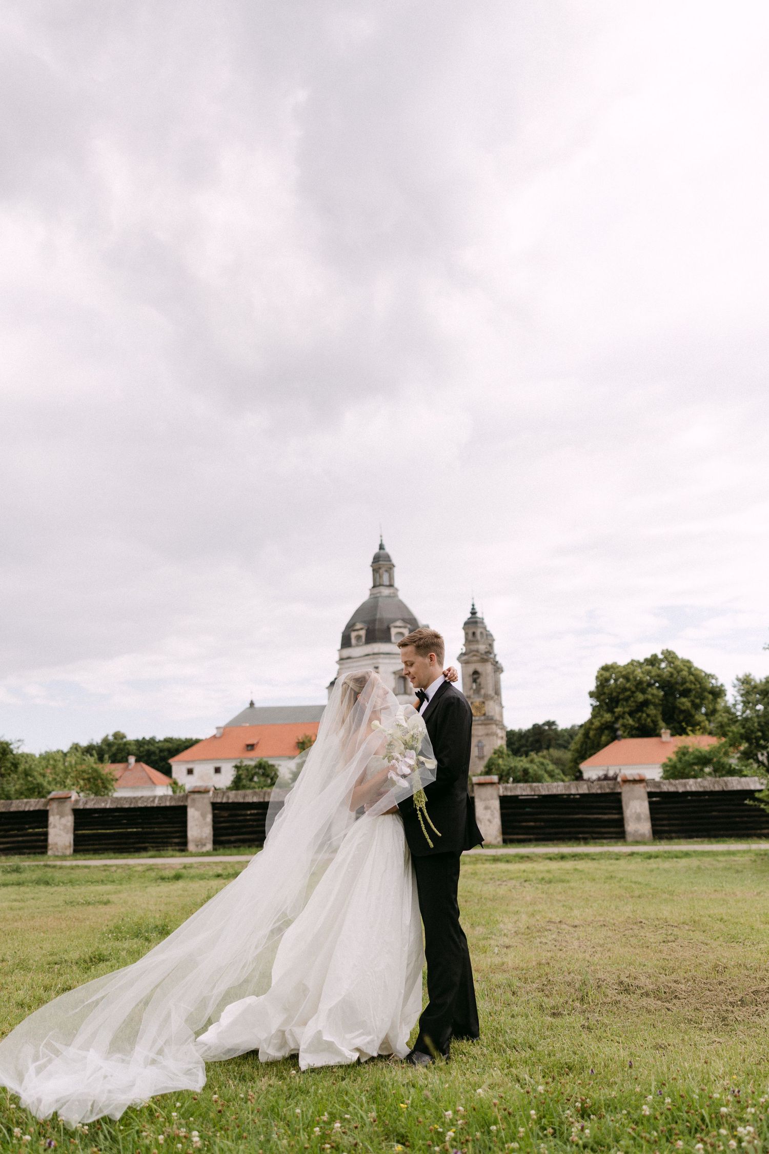 Bride and groom embrace in a meadow with a historic domed church in the background.