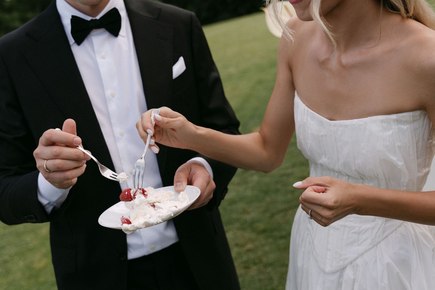 Bride and groom sharing wedding cake outdoors, groom in tuxedo and bride in strapless white gown.